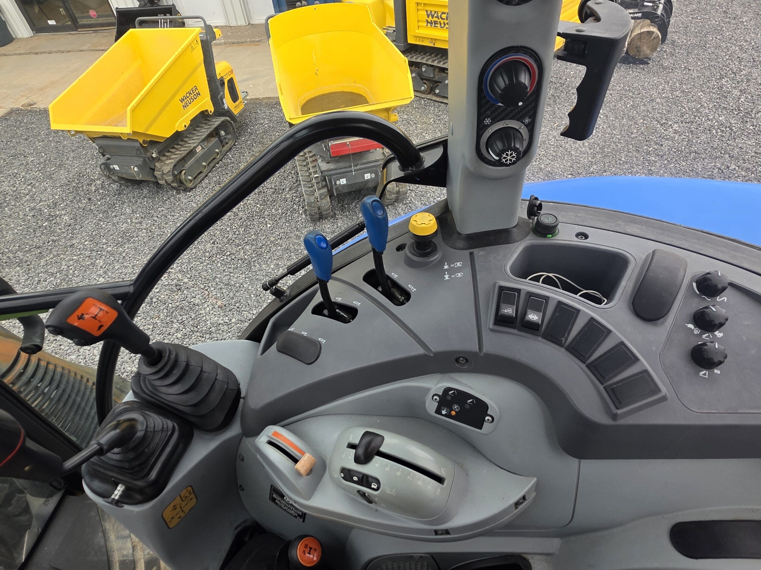 Vue rapprochée du panneau de commande d'un tracteur avec divers leviers, boutons, cadrans et un volant. À l'arrière-plan, deux petits véhicules de construction jaunes sont garés sur du gravier.