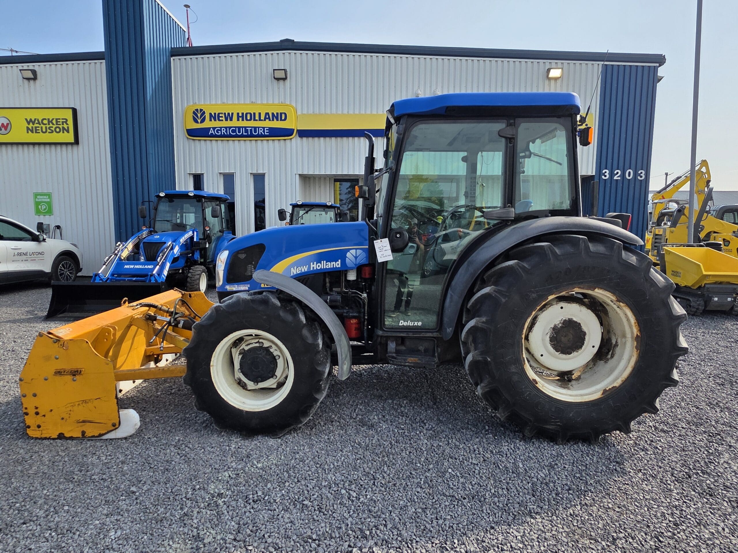 Un tracteur New Holland bleu avec un outil frontal jaune est garé sur du gravier à l'extérieur d'un bâtiment New Holland Agriculture, avec d'autres équipements agricoles et un véhicule blanc visible à l'arrière-plan.
