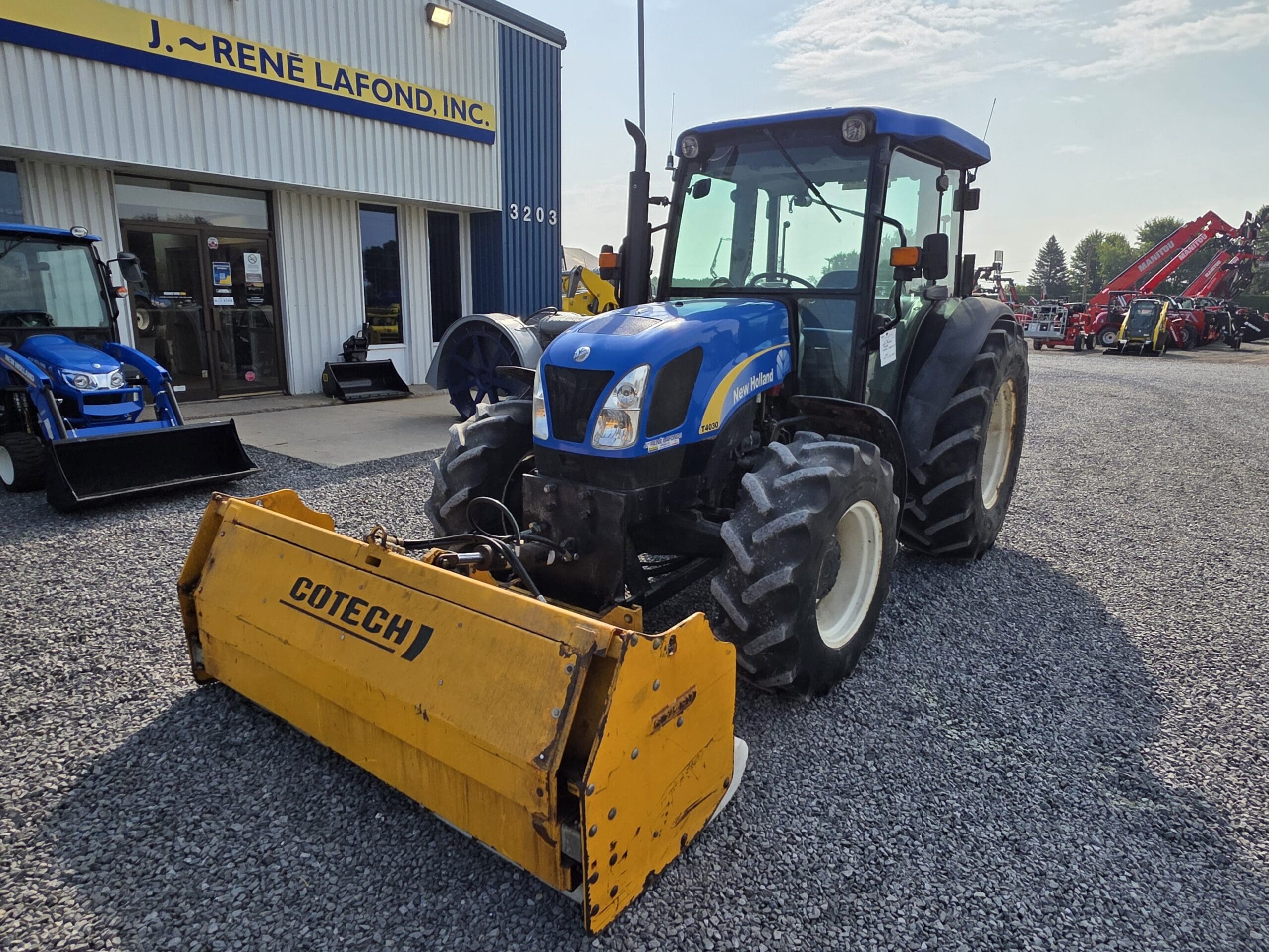Un tracteur New Holland bleu avec un accessoire Cotech jaune est garé sur du gravier à l'extérieur d'un bâtiment portant l'inscription 