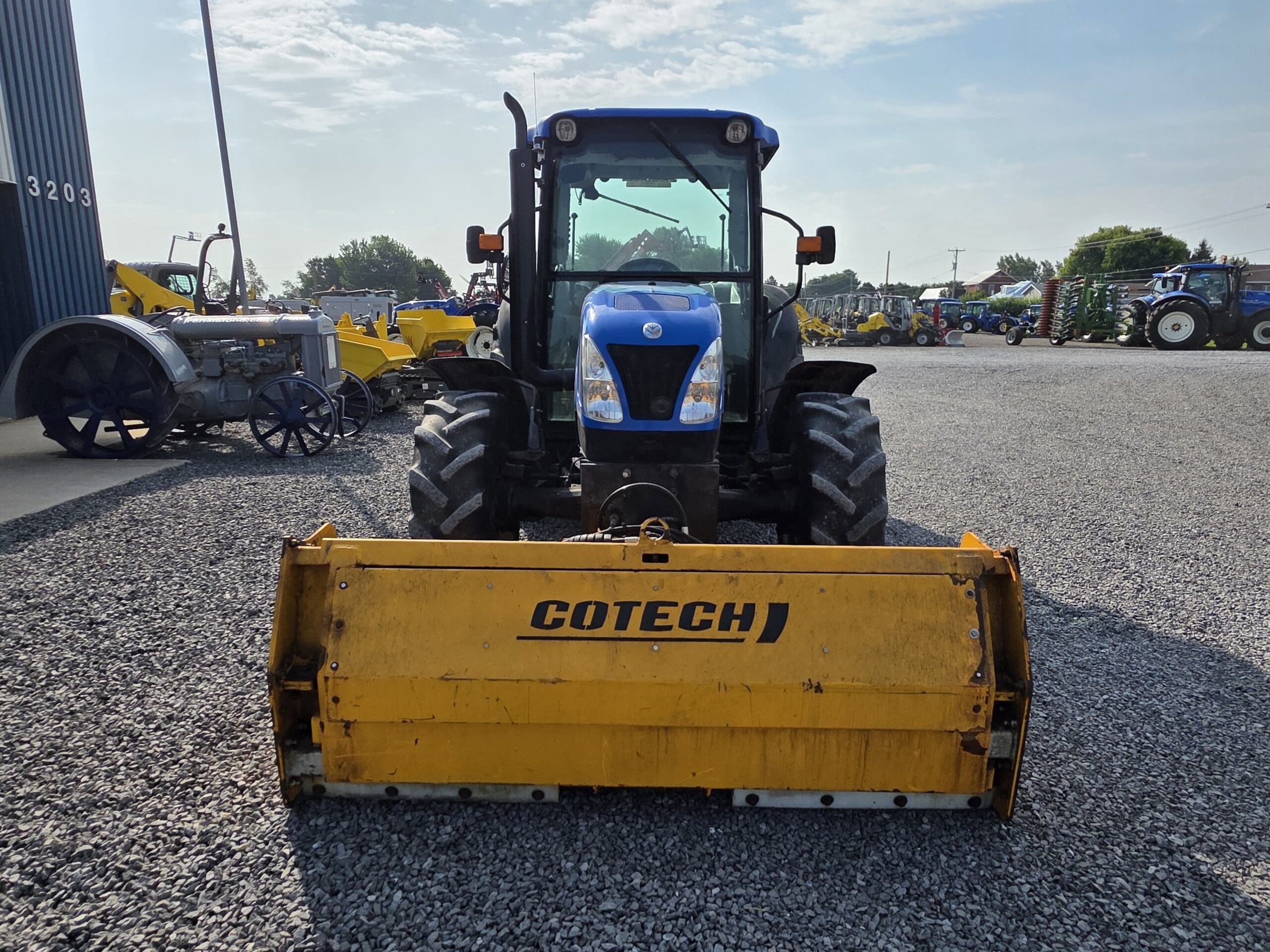 Un tracteur bleu équipé d'un grand accessoire jaune Cotech est garé sur un terrain en gravier. D'autres équipements agricoles sont visibles à l'arrière-plan sous un ciel partiellement nuageux.