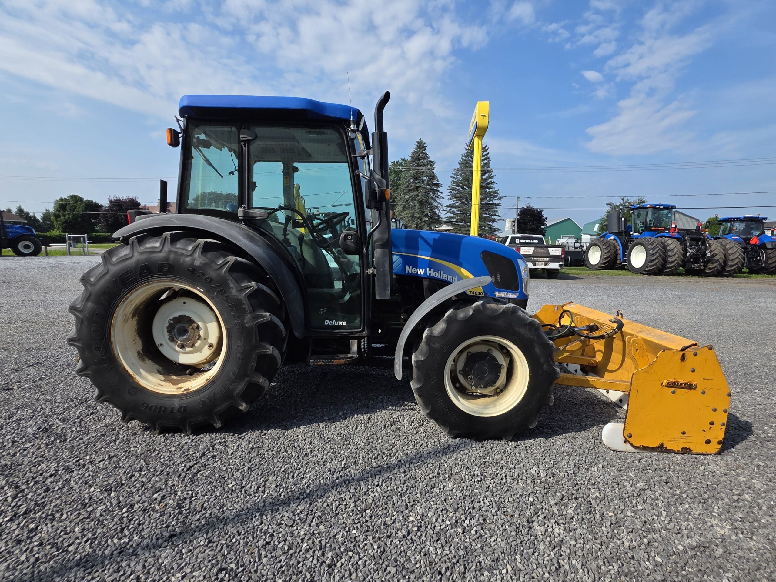 Un tracteur New Holland bleu équipé d'une tondeuse jaune est garé sur un terrain en gravier sous un ciel partiellement nuageux, avec d'autres équipements agricoles visibles à l'arrière-plan.