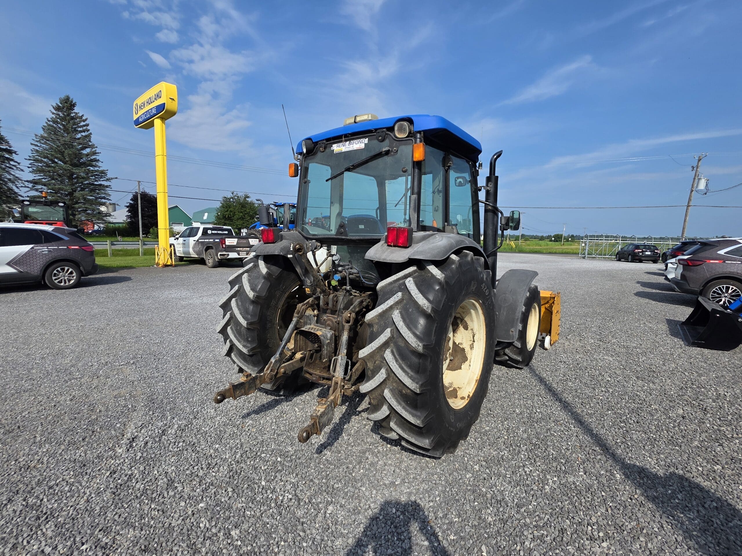 Un tracteur bleu et noir est garé sur un terrain en gravier sous un ciel bleu clair, avec des voitures et une enseigne Leclerc jaune en arrière-plan.