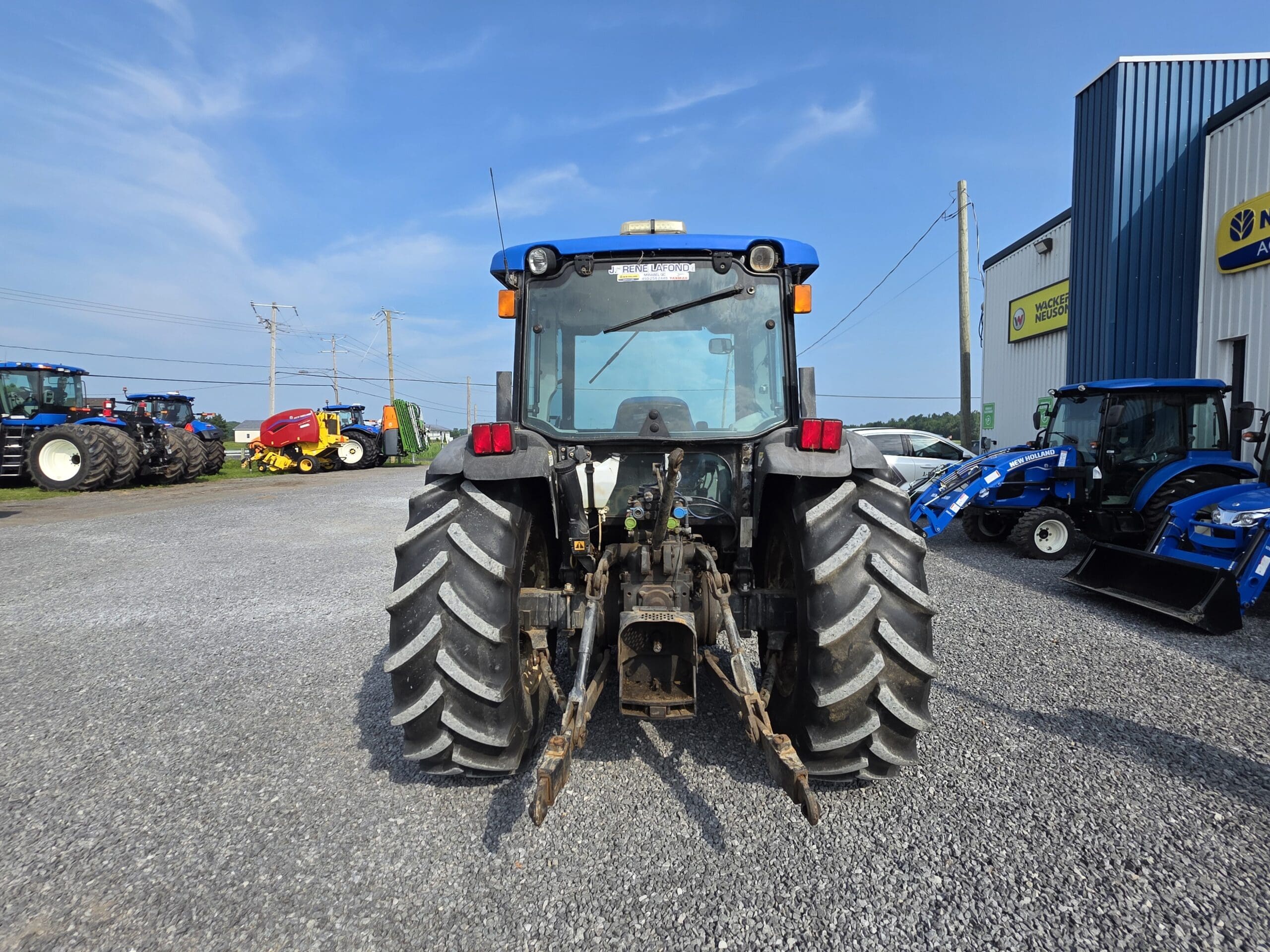 Un tracteur bleu est garé sur du gravier, face à la caméra, avec de gros pneus arrière et du matériel agricole visible à l'arrière-plan, près d'un bâtiment bleu, sous un ciel clair.