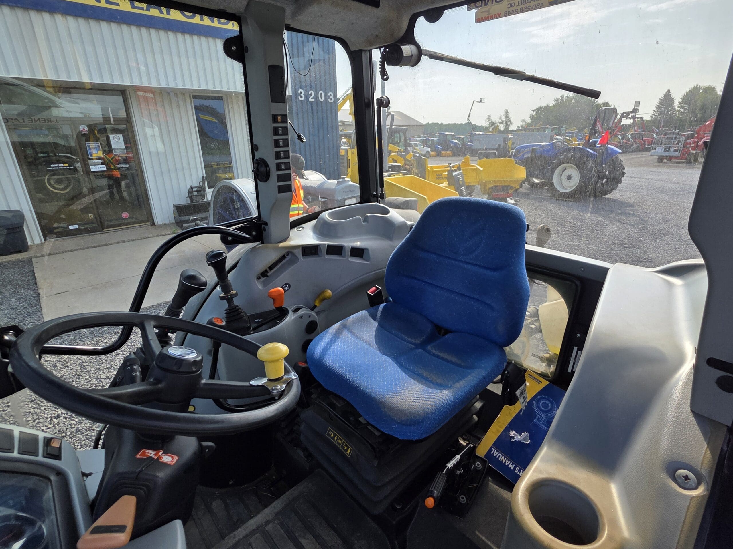 Vue de l'intérieur d'une cabine de tracteur moderne montrant le siège bleu du conducteur, le volant, le panneau de commande et les grandes fenêtres donnant sur d'autres tracteurs et équipements à l'extérieur par une journée ensoleillée.