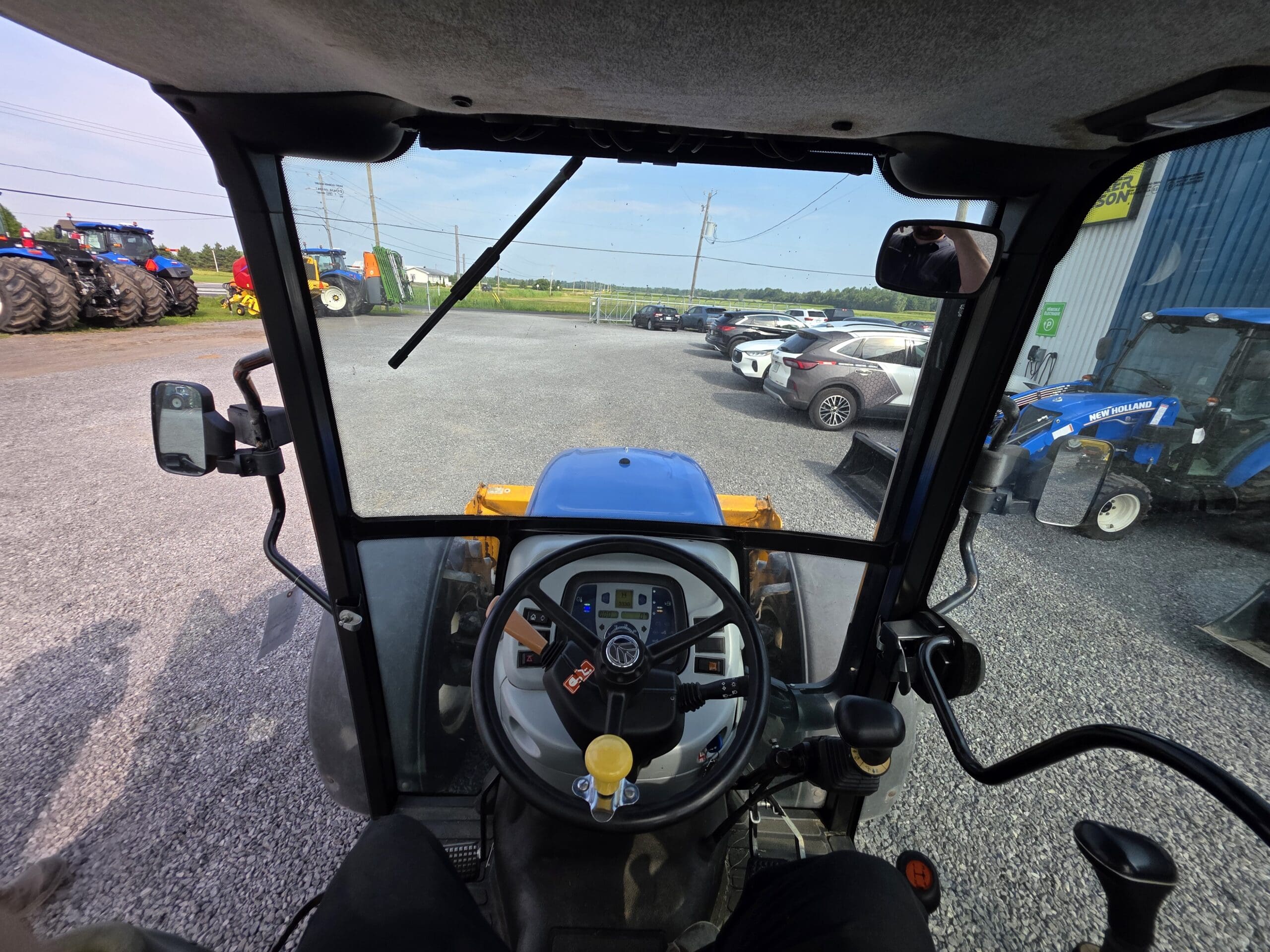 Vue de l'intérieur d'une cabine de tracteur à travers le pare-brise, montrant le volant, les commandes du tableau de bord et les rétroviseurs latéraux. À l'extérieur, plusieurs voitures, tracteurs et équipements agricoles sont visibles dans un parking.