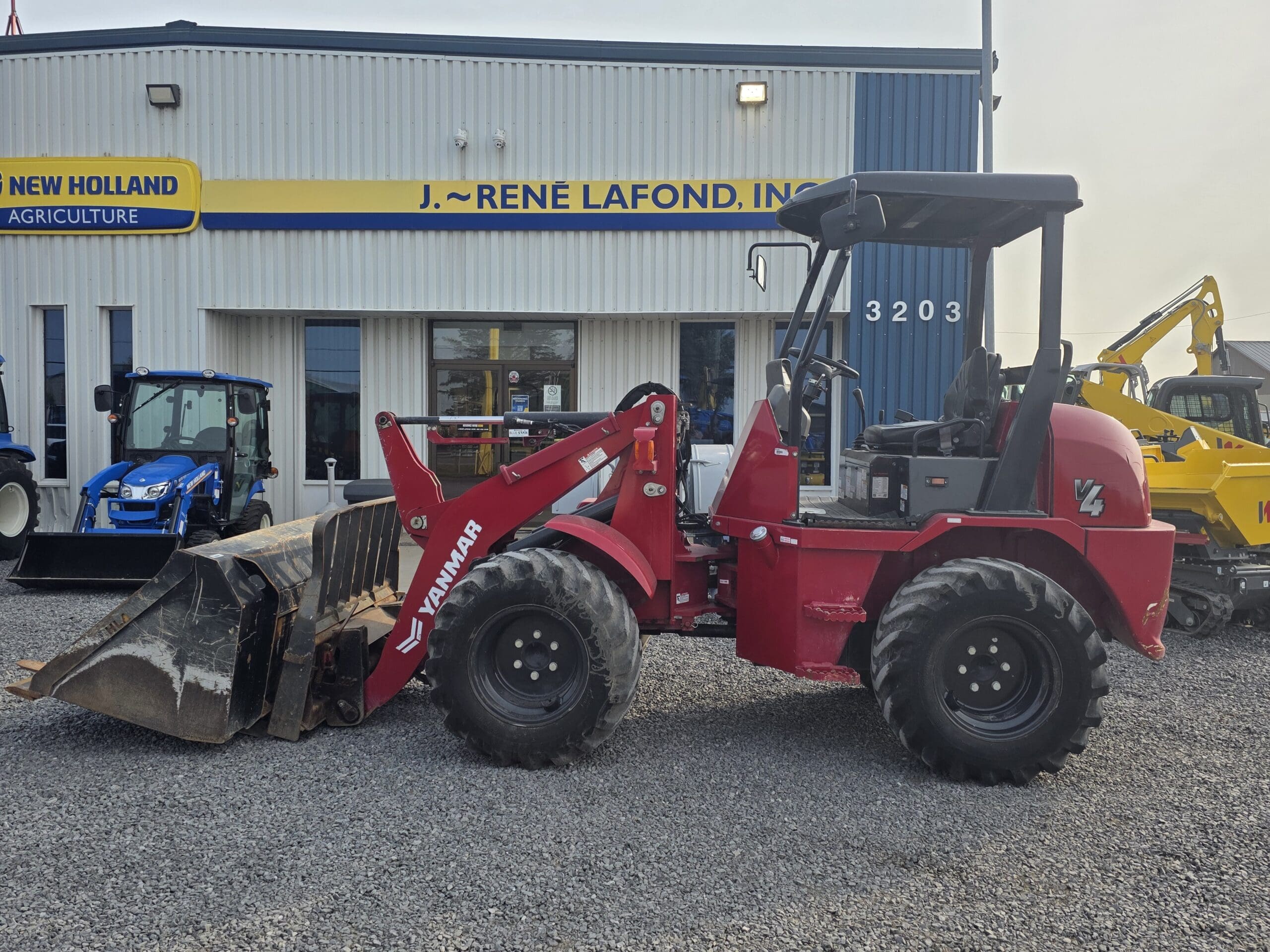 Un chargeur frontal Yanmar rouge équipé d'un grand godet est garé sur du gravier devant un bâtiment portant l'inscription 