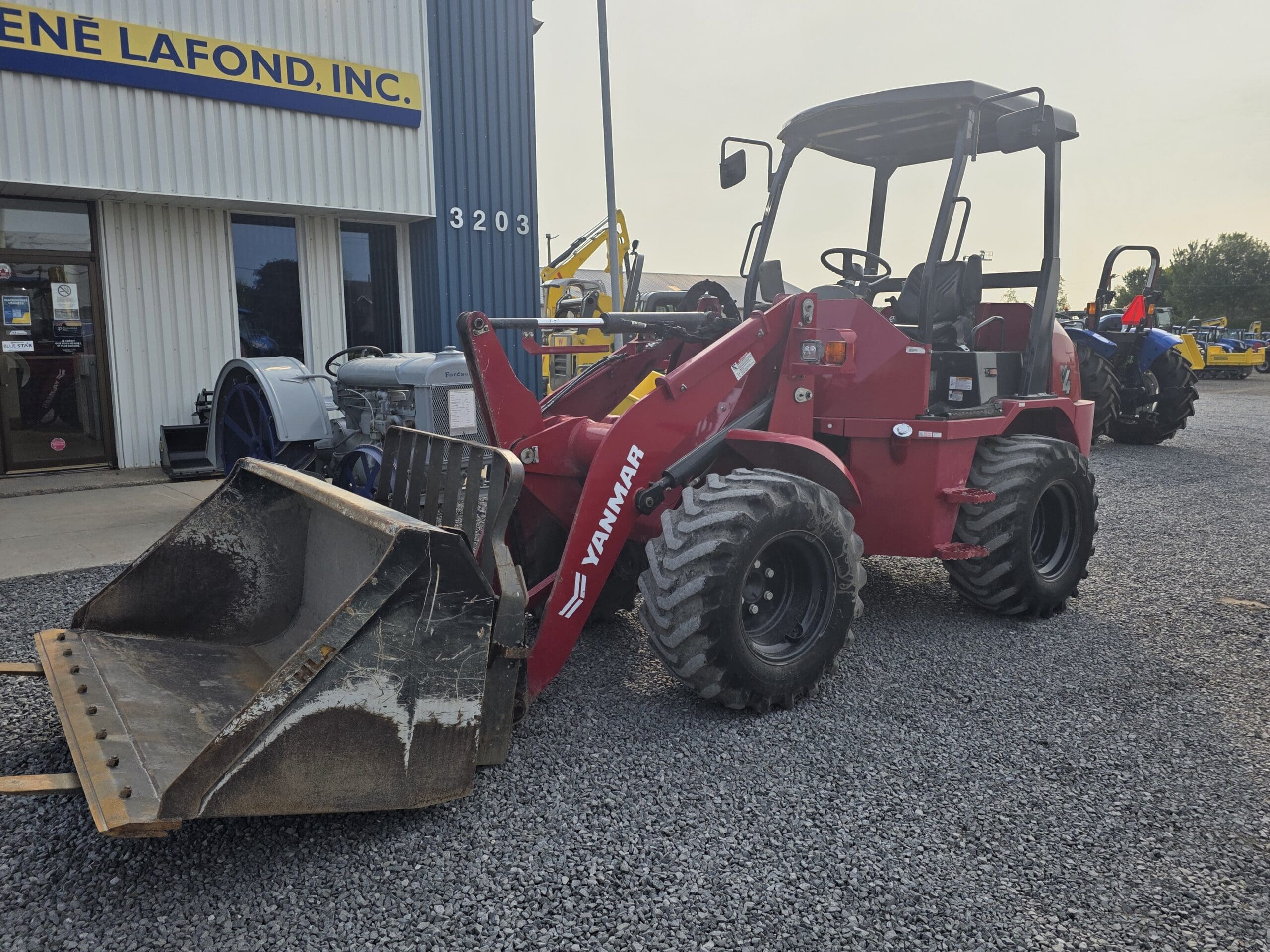Un chargeur frontal Yanmar rouge équipé d'un grand godet est garé sur du gravier à l'extérieur d'un bâtiment portant l'inscription 