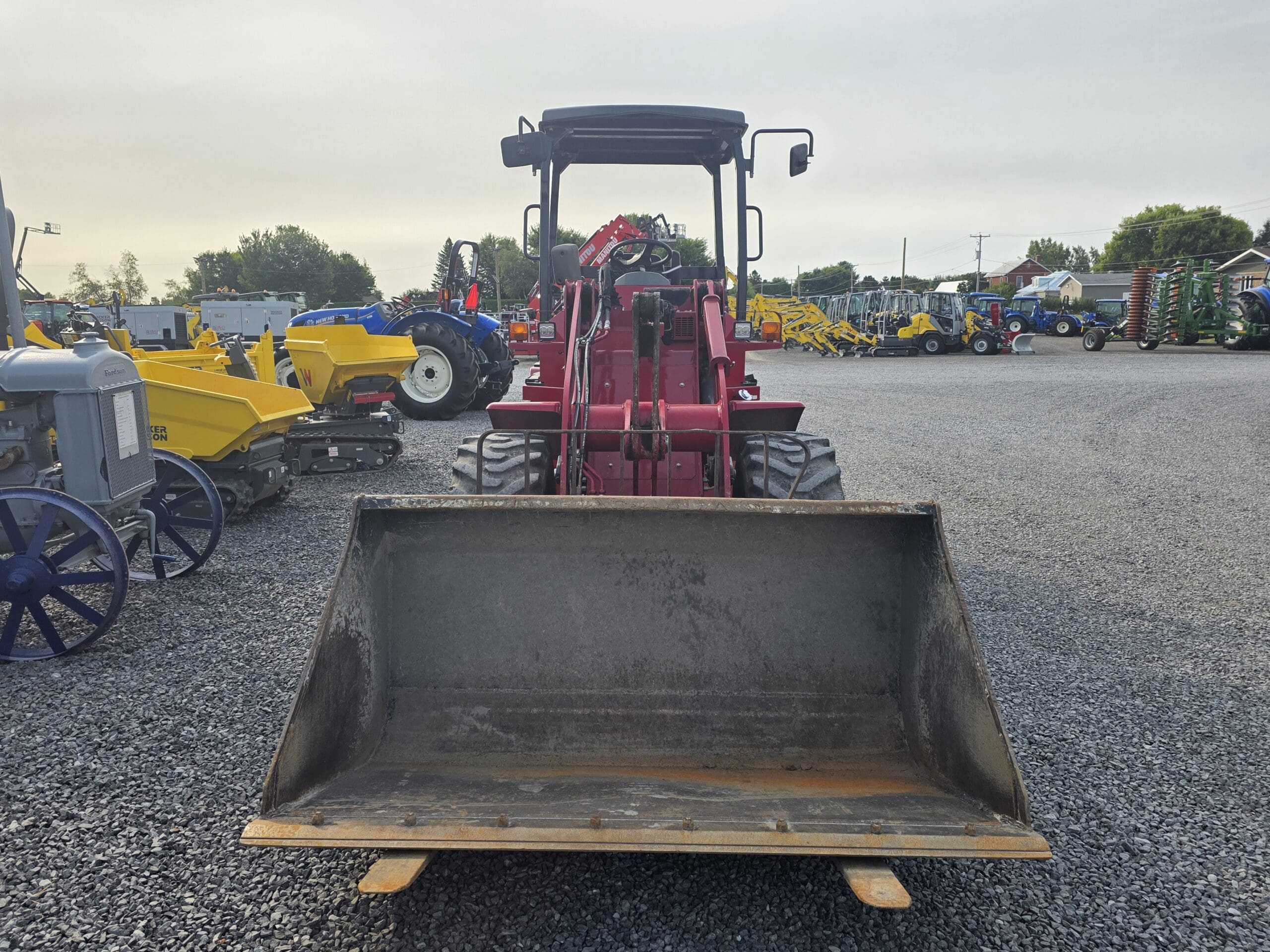 Un tracteur rouge à chargeur frontal est garé face à la caméra dans un terrain en gravier, avec divers autres tracteurs et machines visibles à l'arrière-plan sous un ciel nuageux.