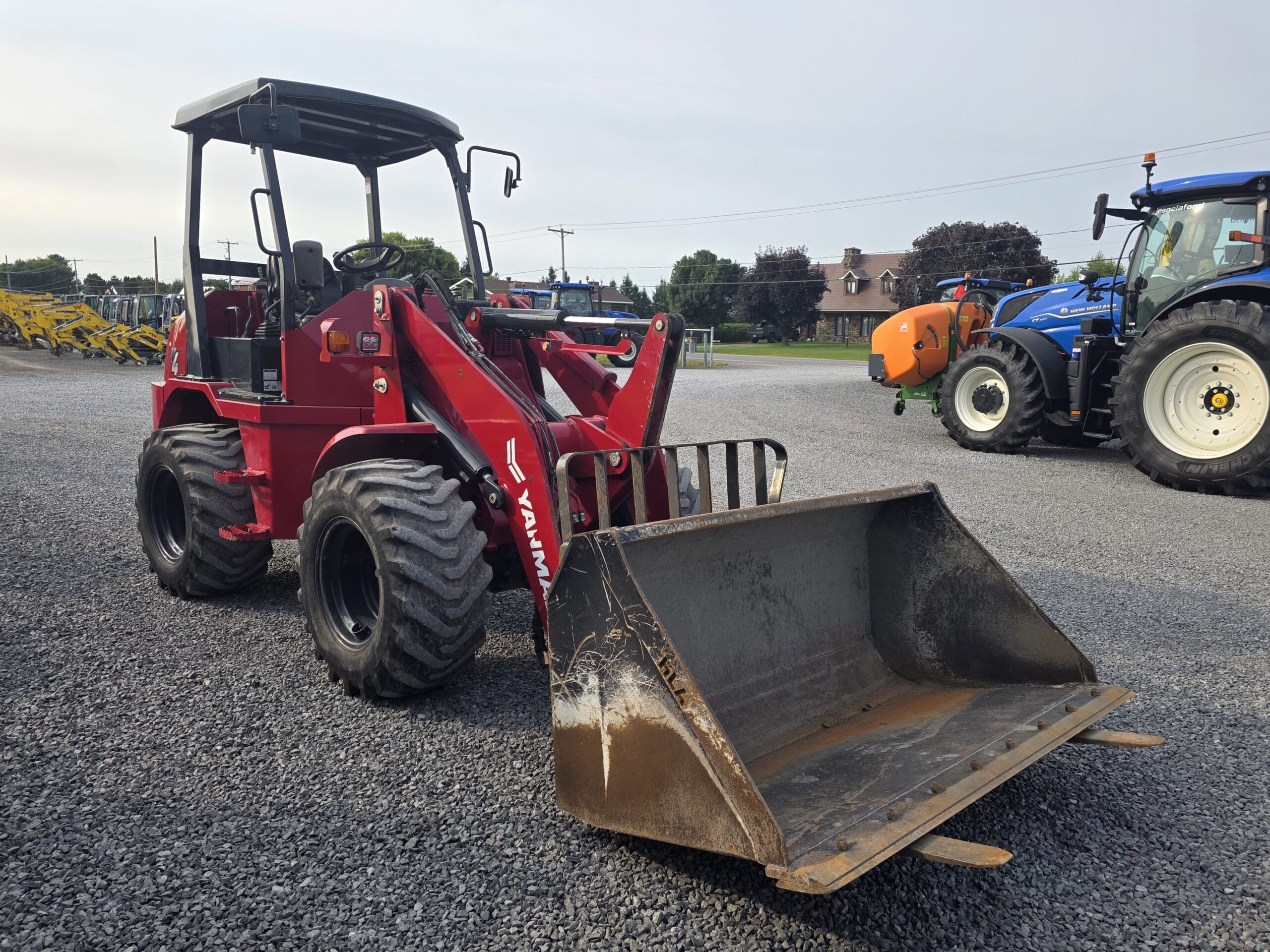 Un chargeur frontal rouge équipé d'un grand godet est garé sur un terrain en gravier. D'autres machines et un tracteur bleu sont visibles à l'arrière-plan sous un ciel nuageux.