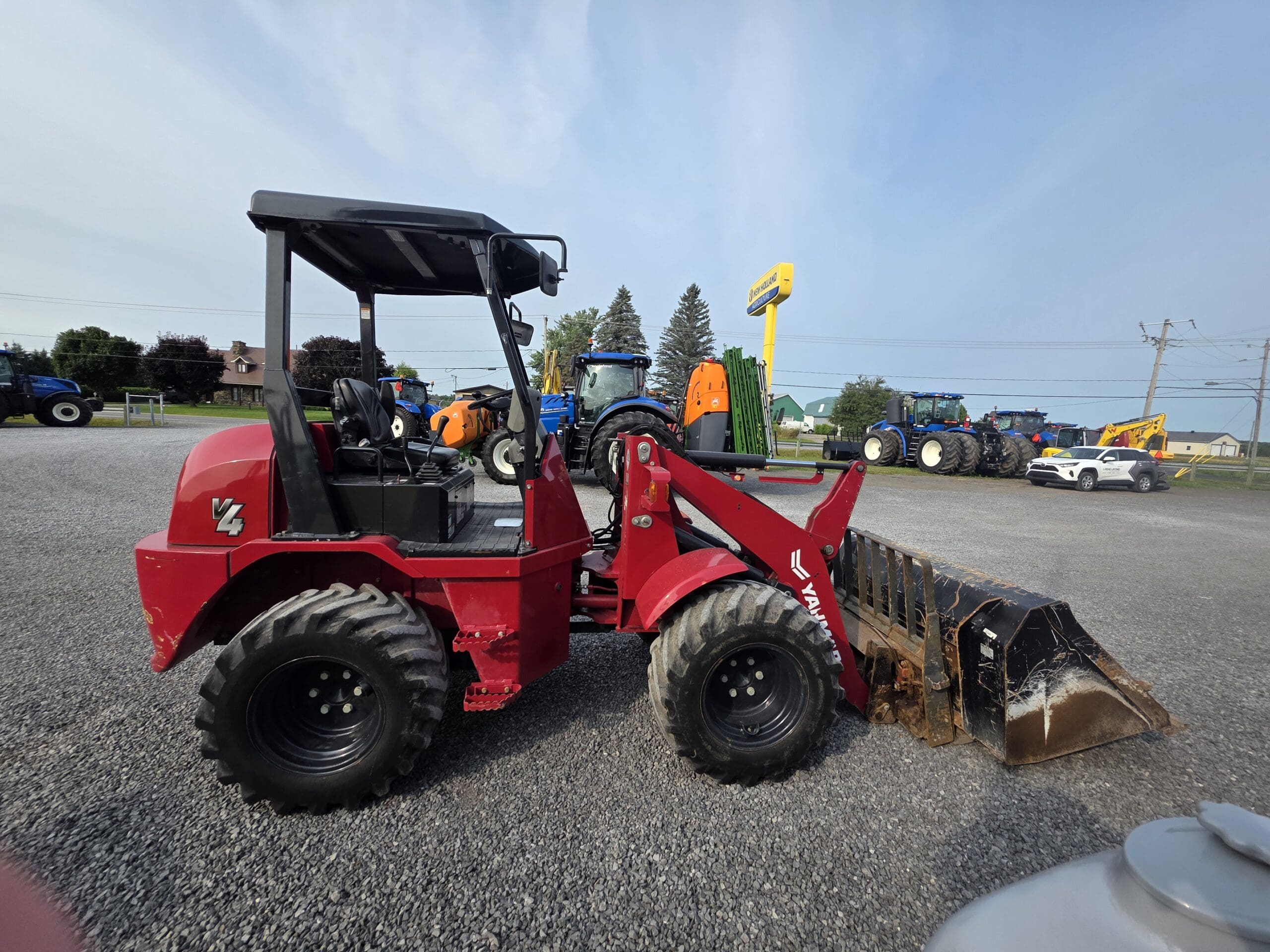 Une chargeuse frontale compacte rouge équipée d'un godet est garée sur du gravier, entourée de divers tracteurs et équipements sous un ciel dégagé.