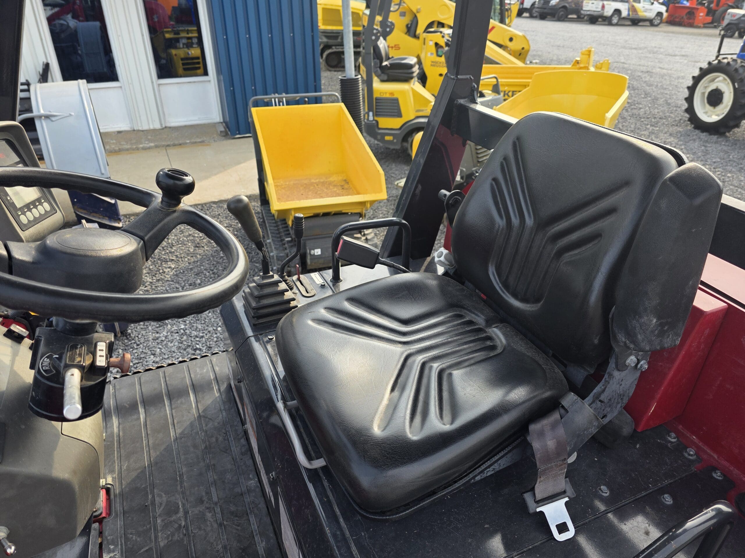 Gros plan d'un siège de tracteur en vinyle noir avec une ceinture de sécurité, un volant et des leviers de commande. À l'arrière-plan, on aperçoit une benne à ordures jaune et divers engins de chantier.