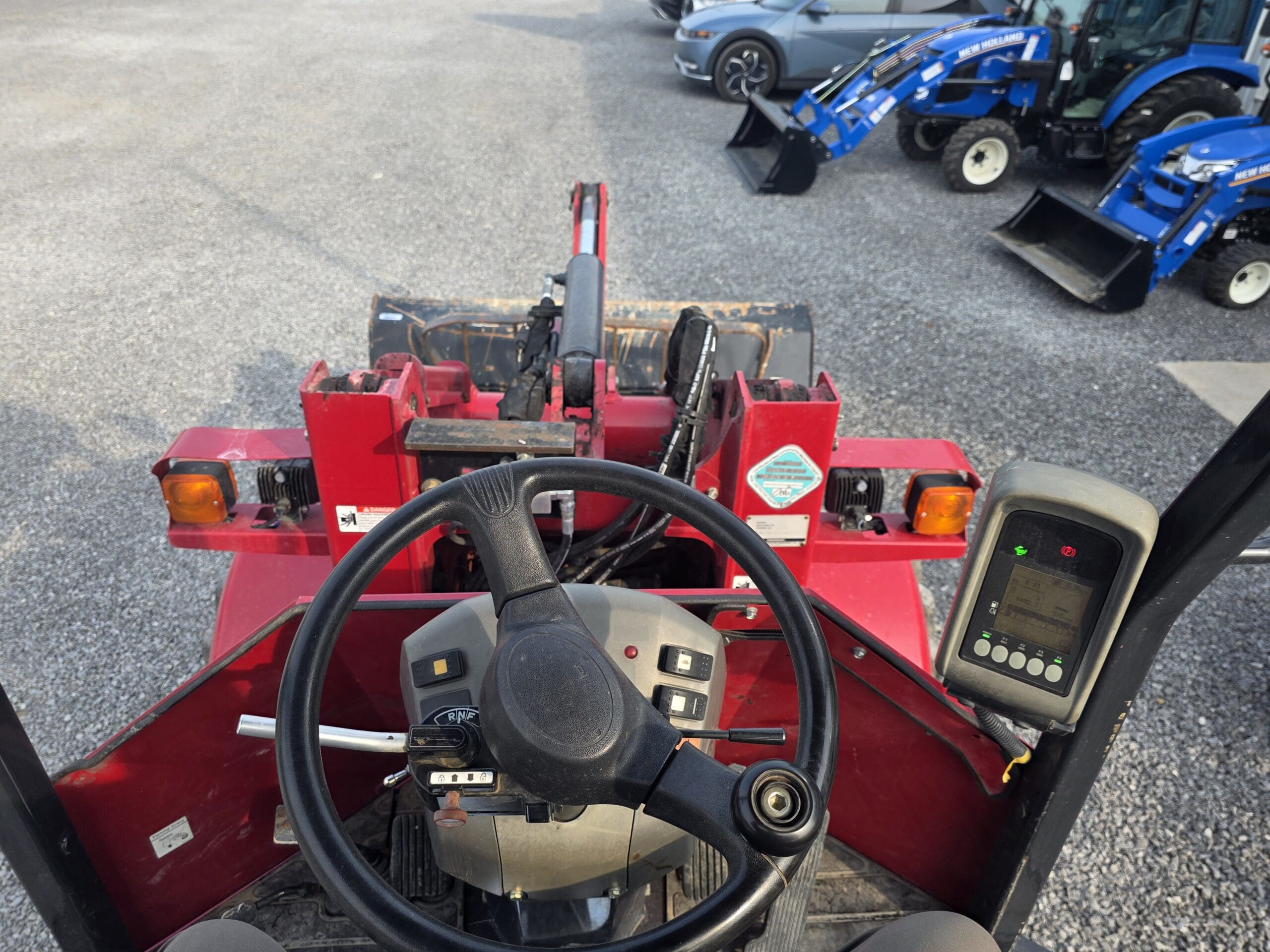 Vue du siège de l'opérateur d'un véhicule à chargeur frontal rouge, montrant le volant, le panneau de commande et l'écran ; des tracteurs bleus et des voitures garées sont visibles à l'arrière-plan sur un terrain en gravier.