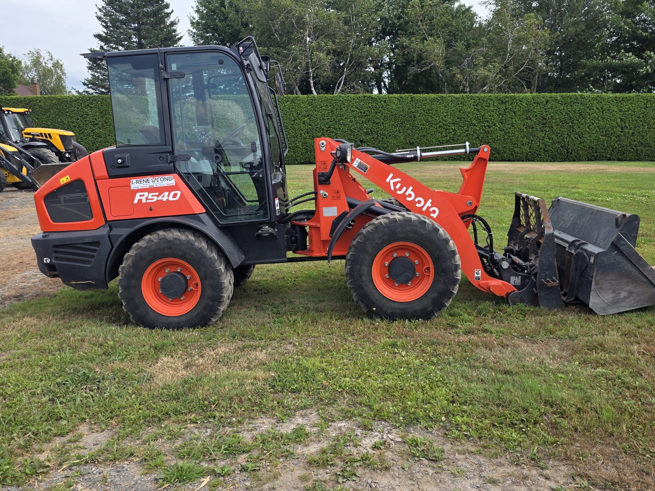 Une chargeuse sur pneus Kubota R540 rouge équipée d'un godet avant est garée sur de l'herbe près d'une haie et d'arbres.