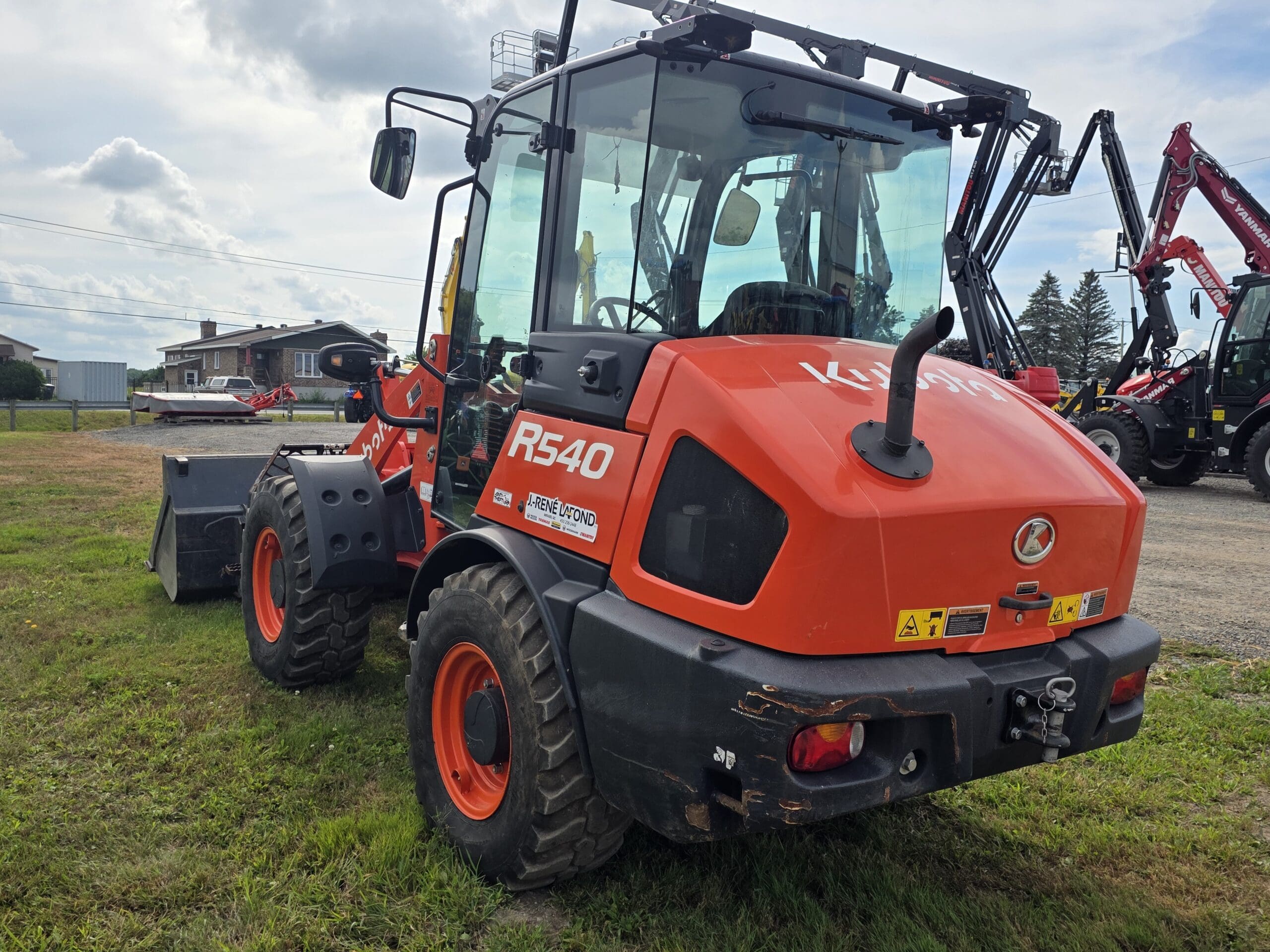 Une chargeuse compacte sur pneus Kubota R540 orange vif est garée sur de l'herbe avec d'autres machines en arrière-plan sous un ciel nuageux. La chargeuse est vue de l'arrière gauche.