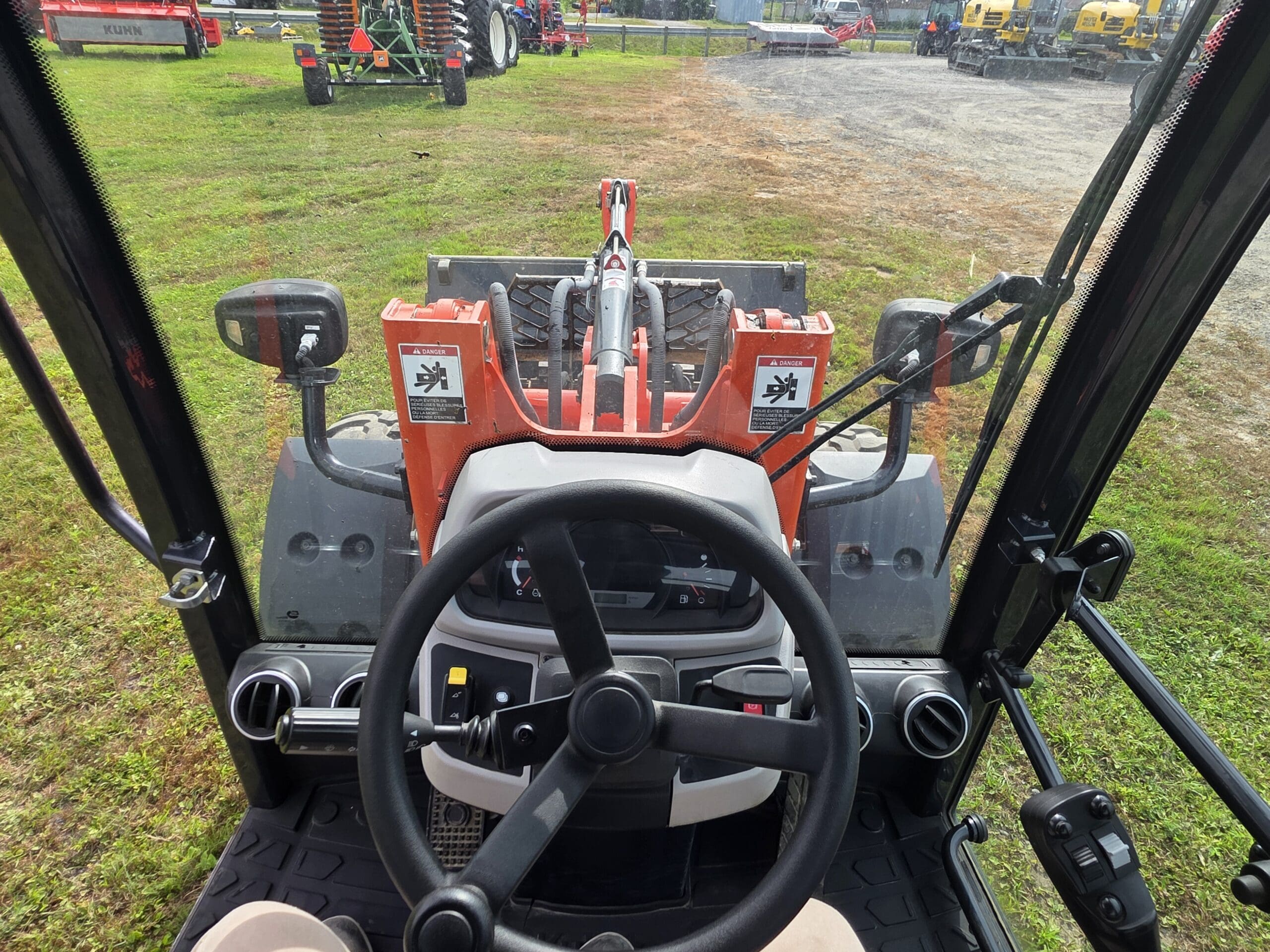 Vue de l'intérieur de la cabine d'un tracteur, montrant le volant, les commandes et le chargeur frontal à l'extérieur, avec de l'herbe et d'autres équipements agricoles visibles à l'arrière-plan.