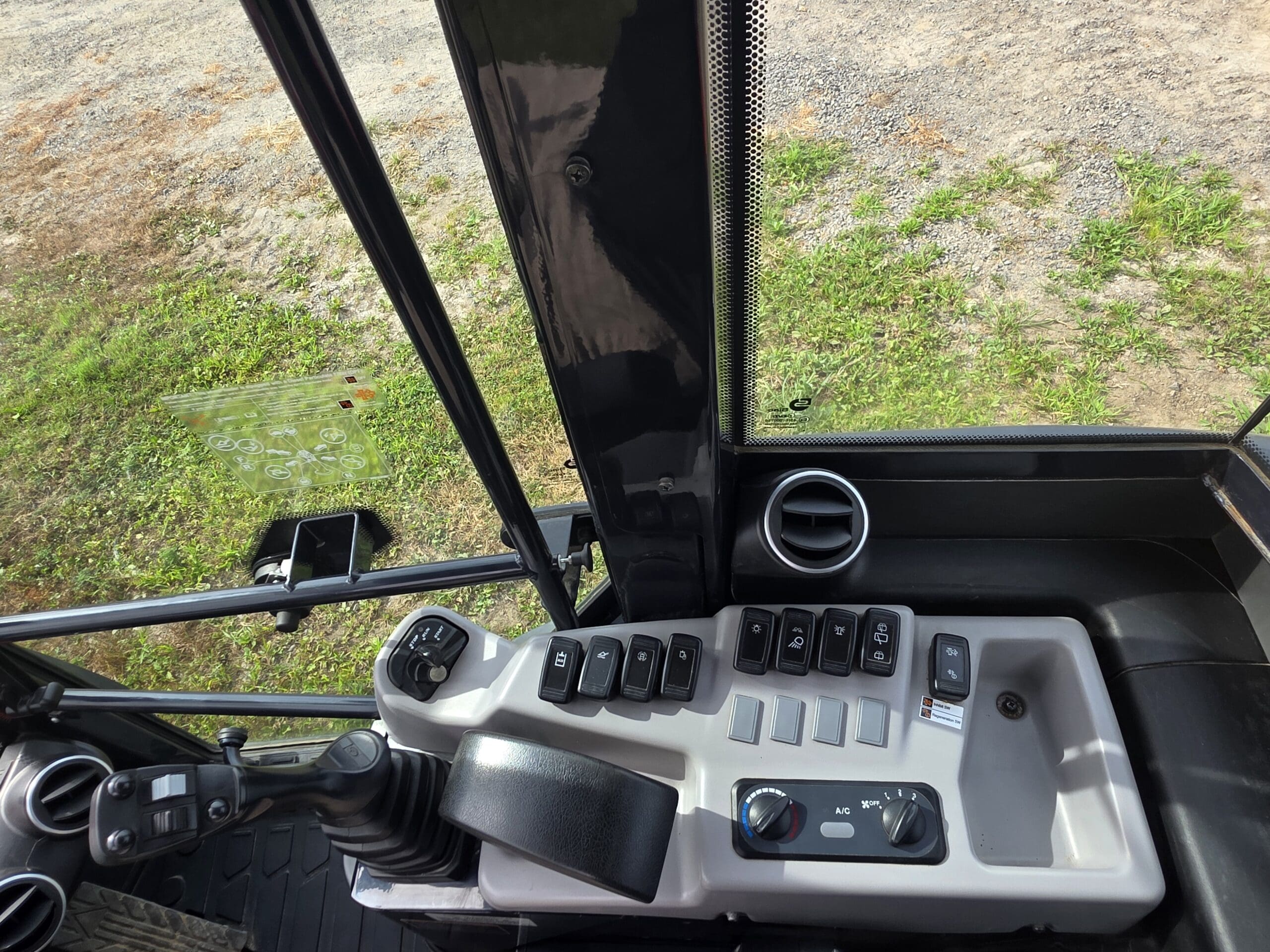 Panneau de commande d'un véhicule de chantier, montrant plusieurs interrupteurs, des cadrans et un joystick. La vue comprend une partie du siège et de la fenêtre du conducteur, avec du gravier et de l'herbe visibles à l'extérieur.