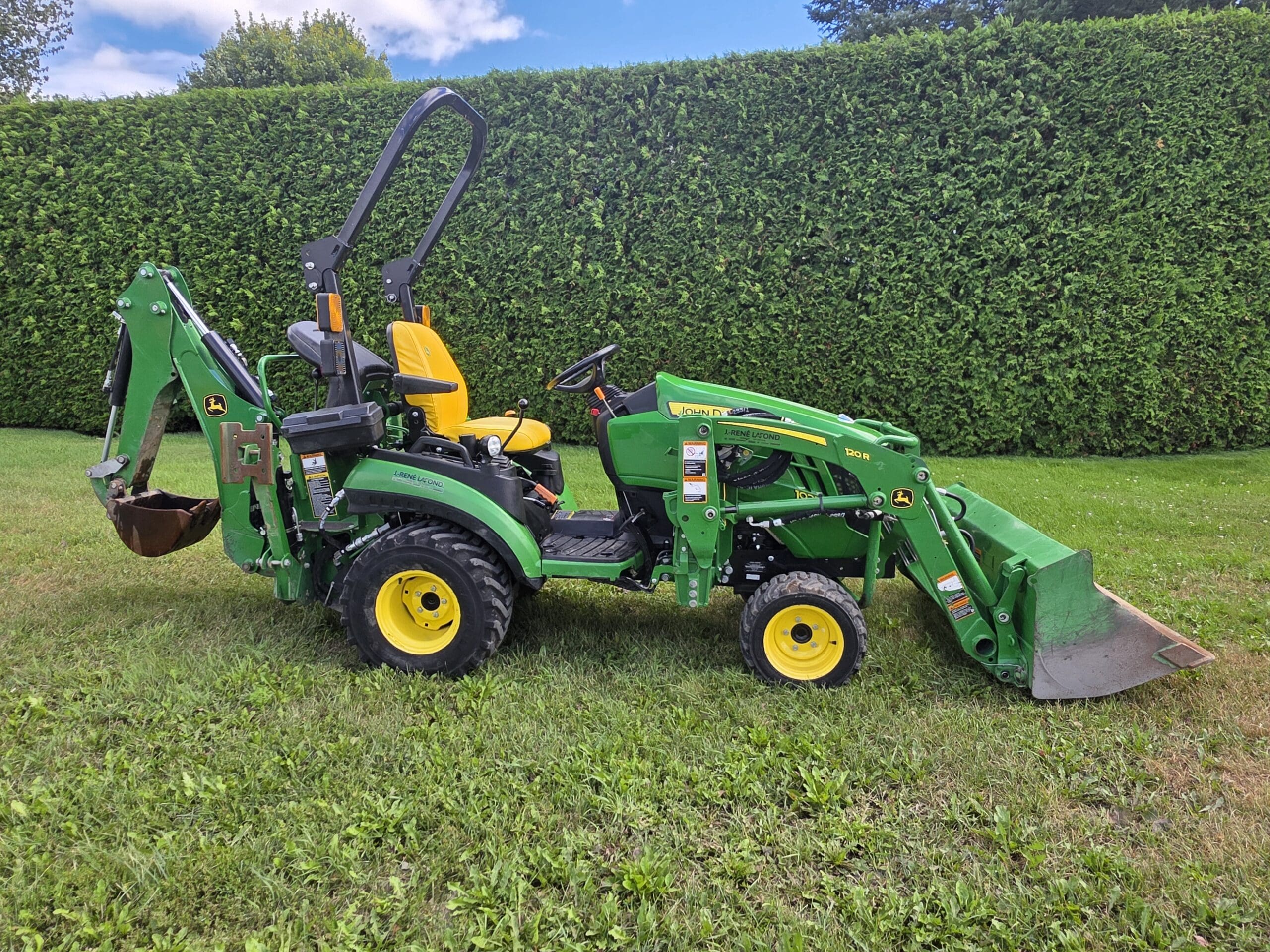 Un tracteur compact vert et jaune, équipé d'un chargeur frontal et d'une pelleteuse, est posé sur l'herbe devant une haie verte haute et dense.
