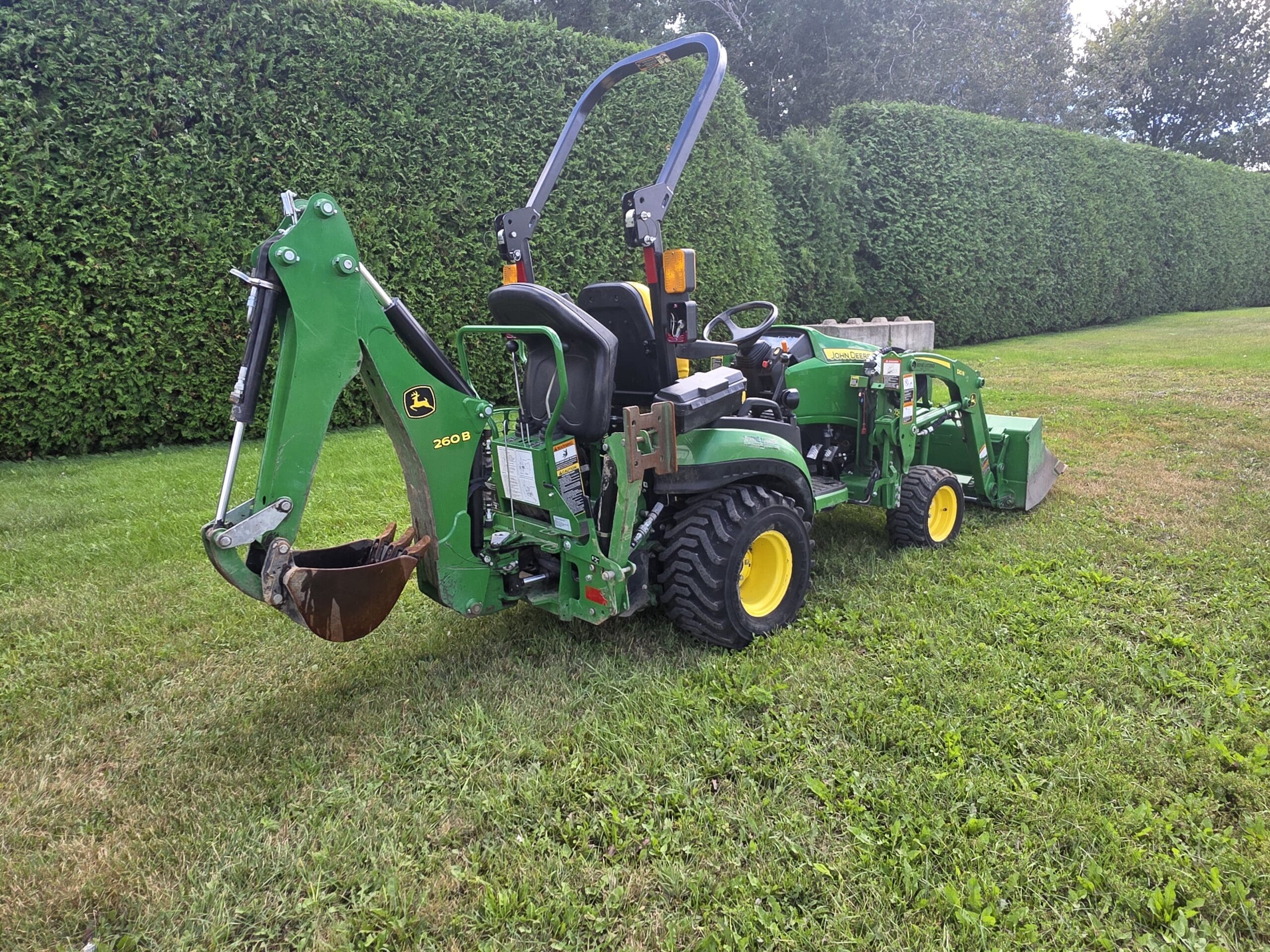 Un petit tracteur vert John Deere équipé d'une pelleteuse et d'un chargeur frontal est garé sur une pelouse herbeuse à côté d'une haie haute et bien taillée à l'arrière-plan.