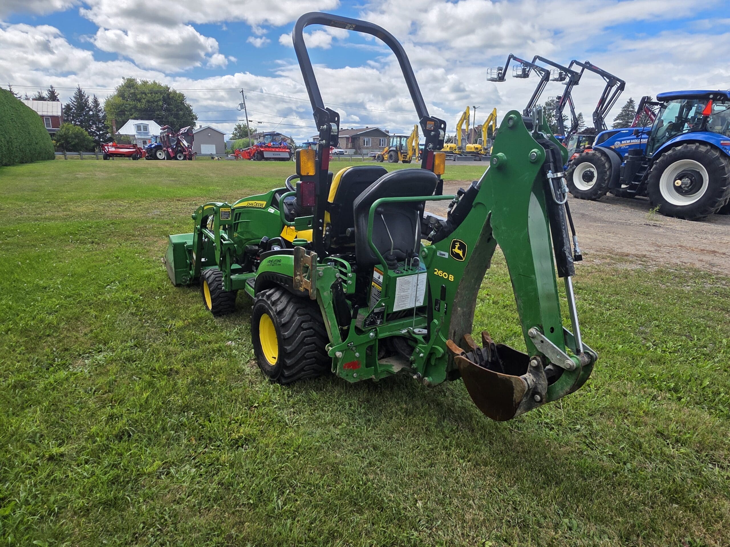 Un tracteur pelleteuse John Deere vert est garé sur de l'herbe, avec divers autres tracteurs et machines visibles en arrière-plan, sous un ciel partiellement nuageux.