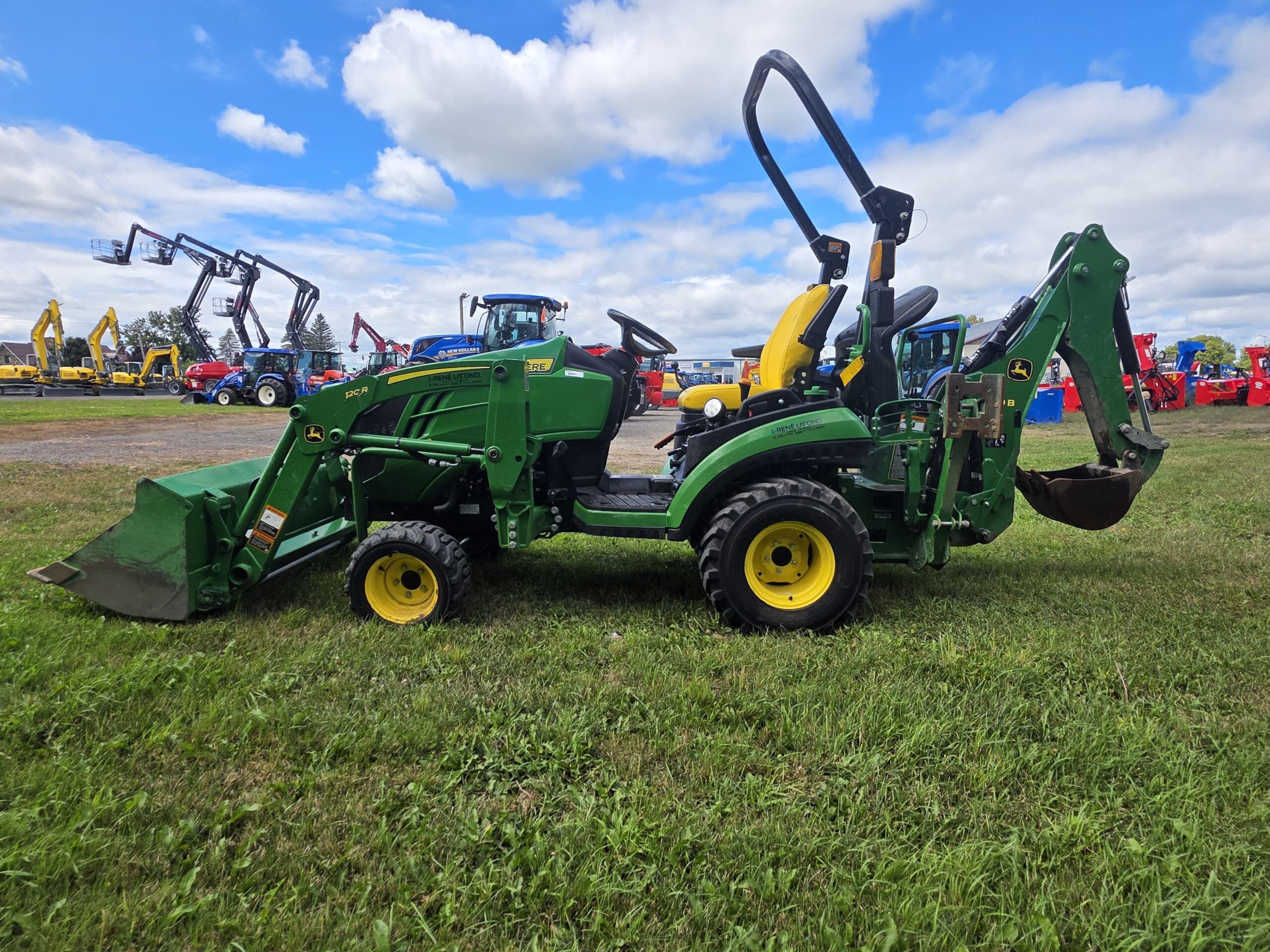 Un tracteur vert et jaune équipé d'un chargeur frontal et d'une pelleteuse est posé sur de l'herbe sous un ciel bleu partiellement nuageux. D'autres tracteurs et machines sont visibles à l'arrière-plan.