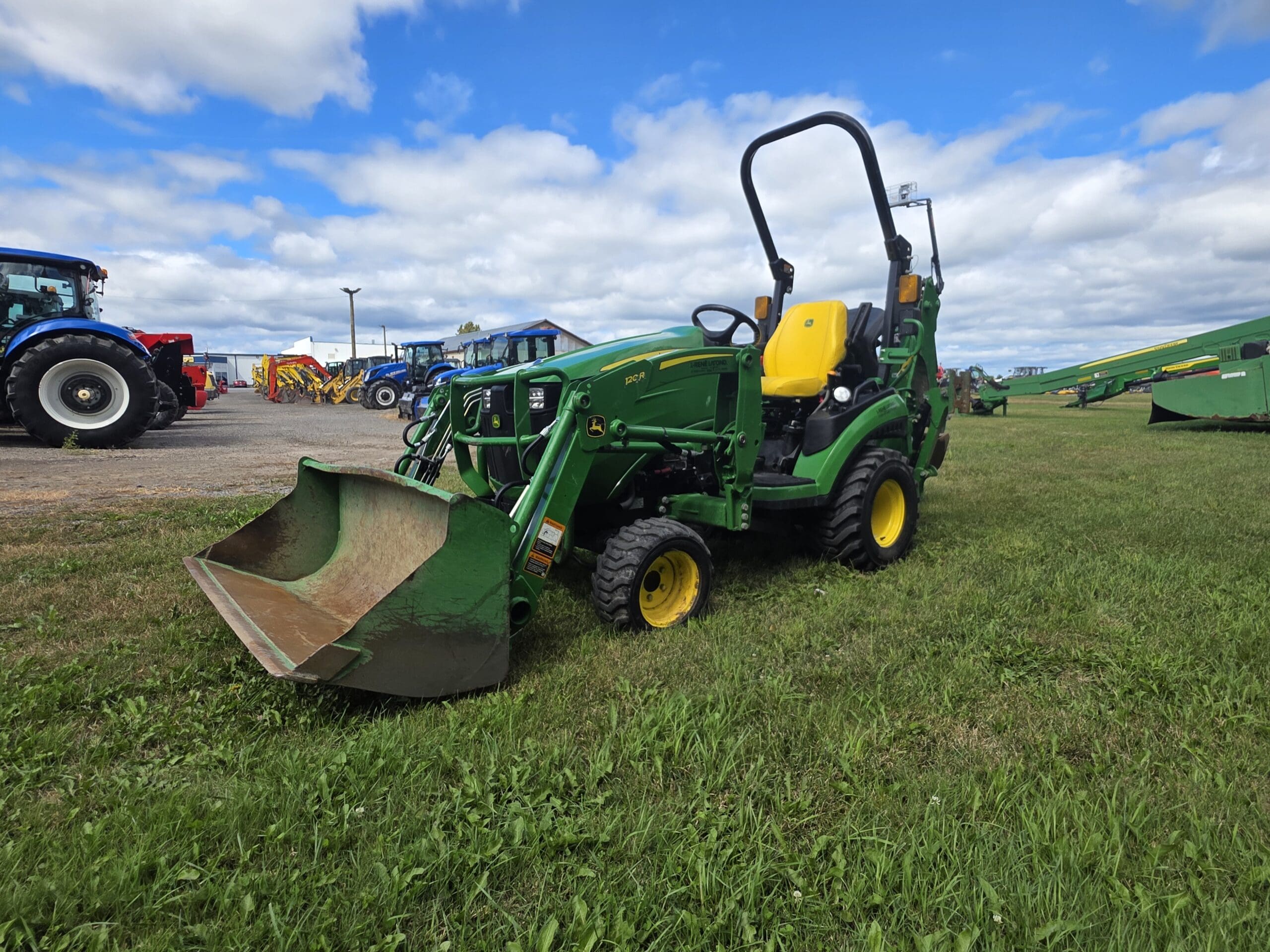 Un tracteur vert et jaune équipé d'un chargeur frontal est posé sur de l'herbe sous un ciel partiellement nuageux. D'autres équipements agricoles et tracteurs sont visibles à l'arrière-plan.
