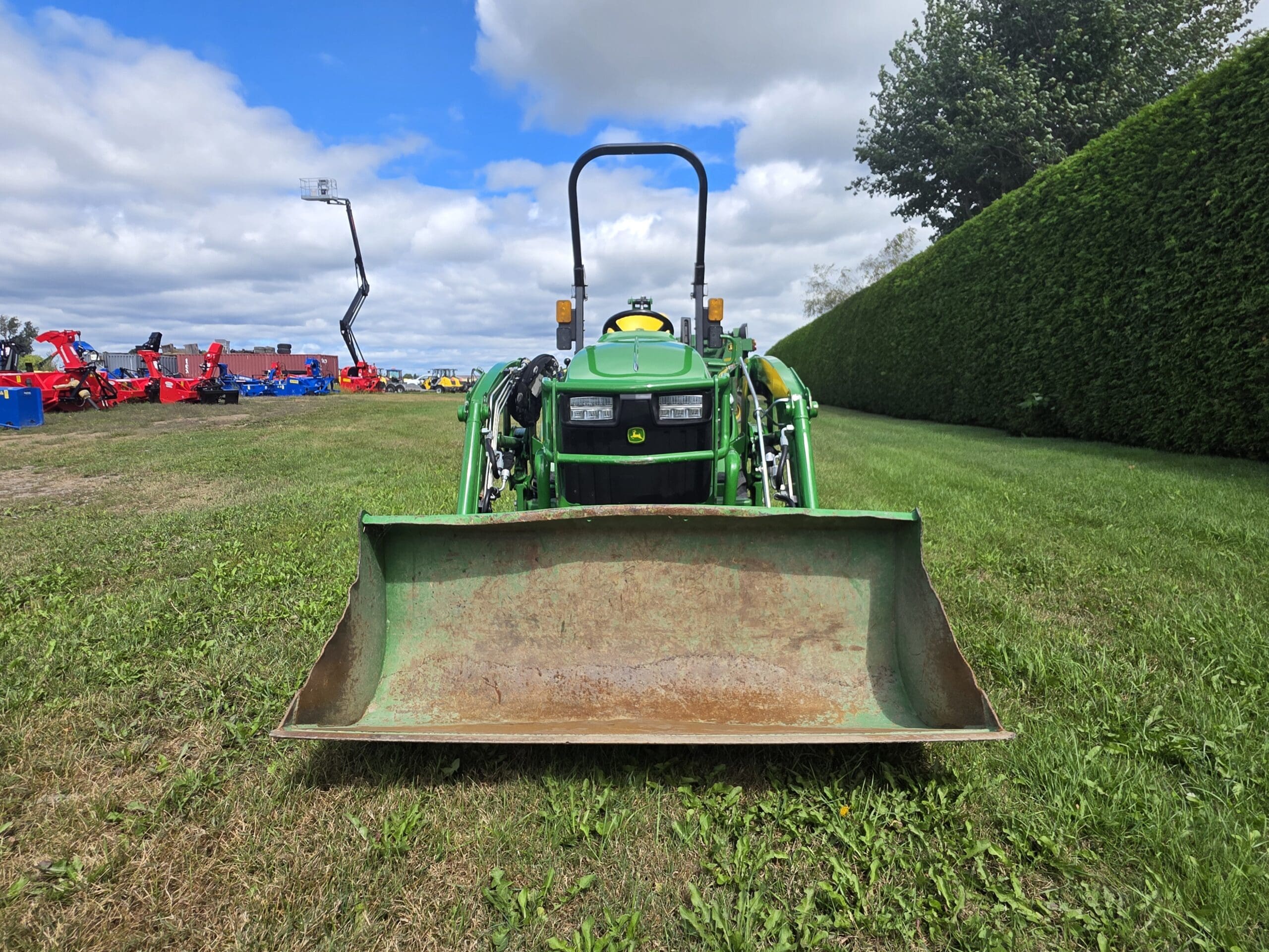 Un tracteur vert équipé d'un chargeur frontal est garé sur de l'herbe, face à la caméra. À l'arrière-plan, on aperçoit d'autres machines agricoles et des arbres sous un ciel partiellement nuageux.
