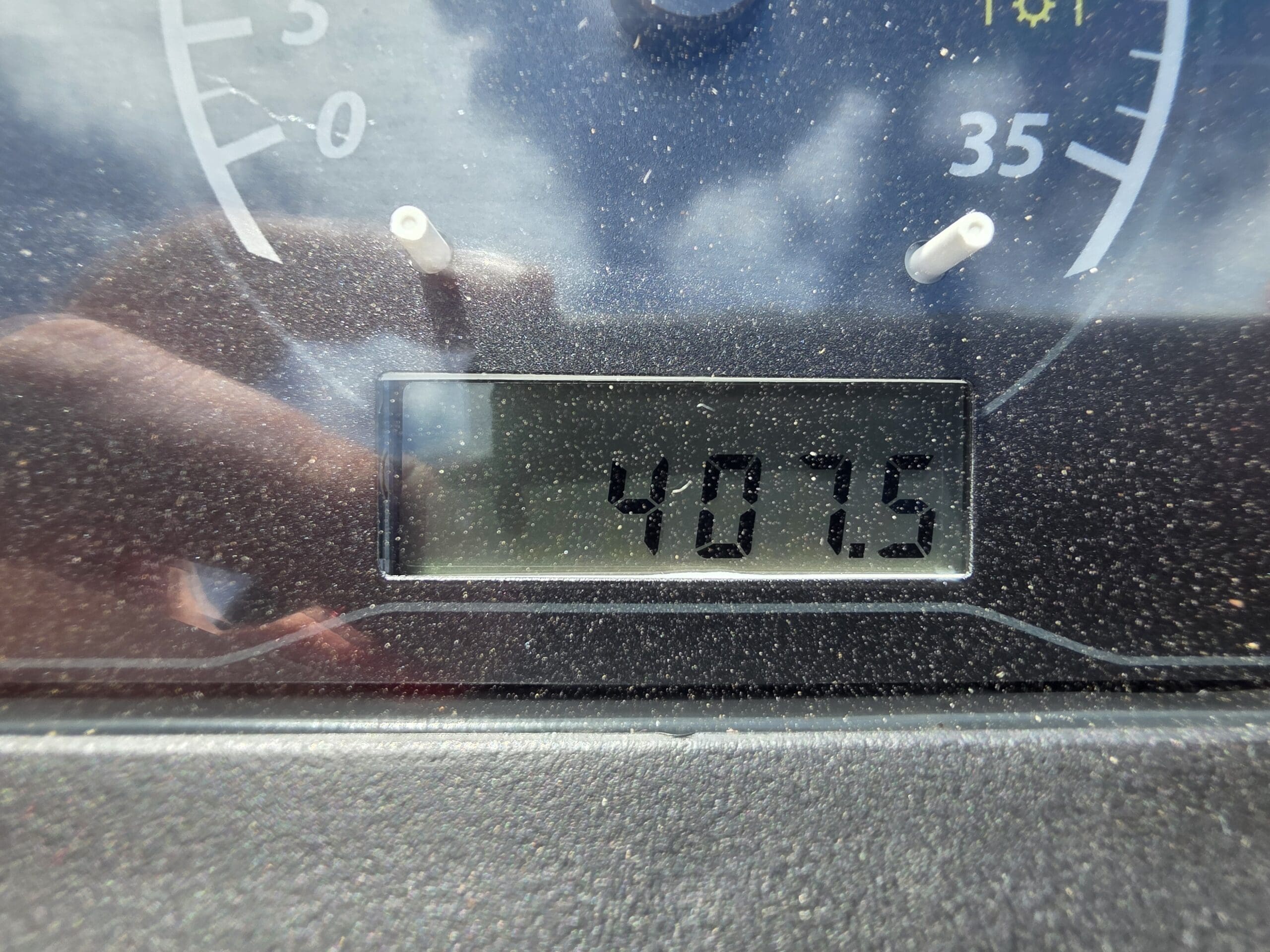 Gros plan de l'affichage numérique poussiéreux du compteur kilométrique d'un véhicule indiquant le nombre 4075, avec une partie d'une jauge analogique et un faible reflet visible sur la vitre.
