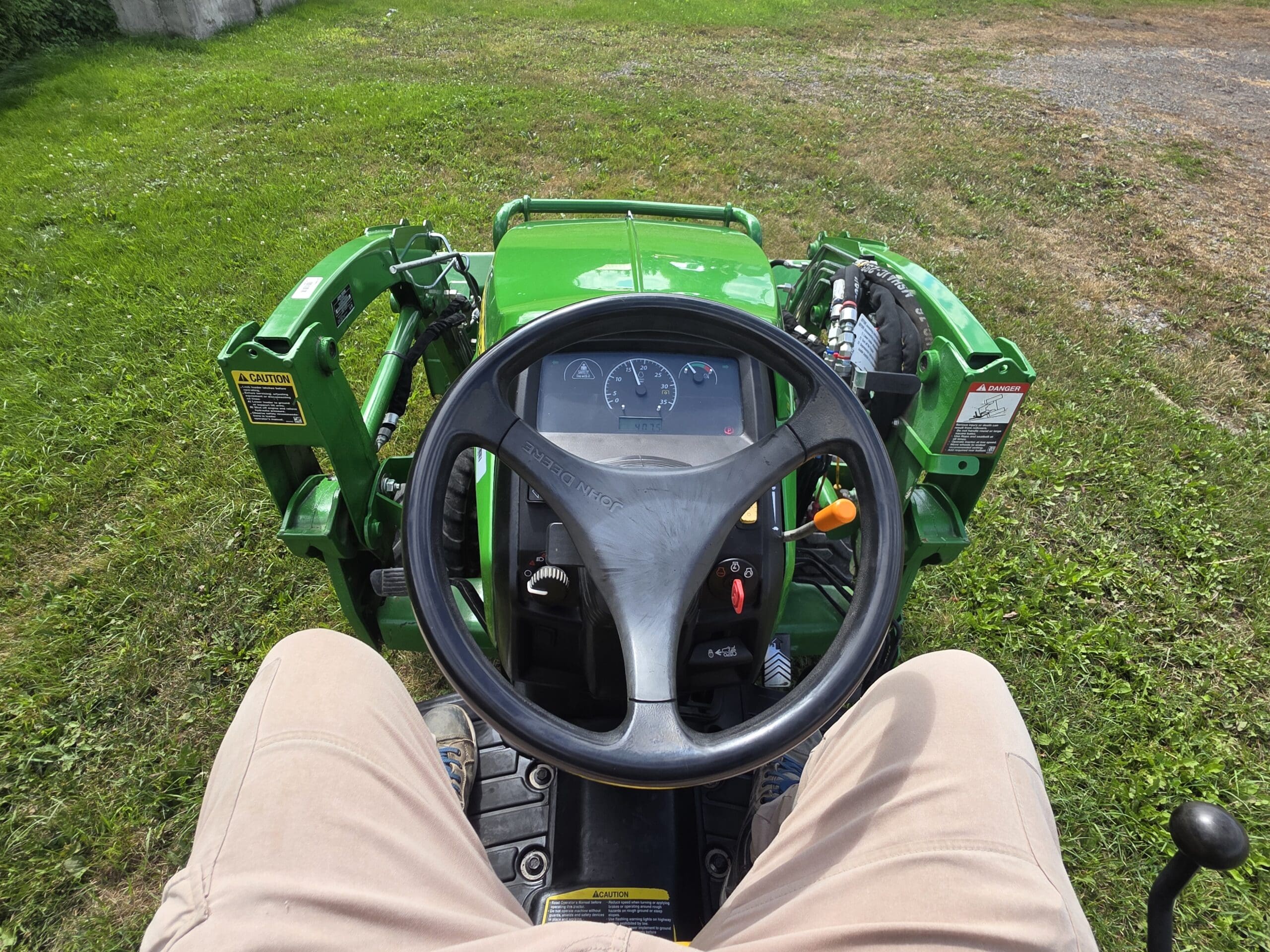 Vue d'une personne depuis le siège du conducteur d'un tracteur vert, montrant le volant, le panneau de commande et ses jambes, avec de l'herbe et un chemin de gravier visibles à l'extérieur.