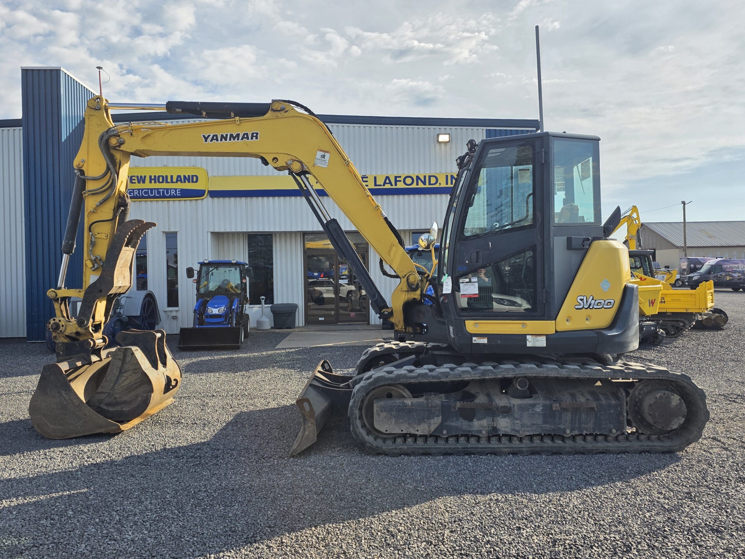 Une pelle compacte Yanmar SV40 jaune avec un godet est garée sur du gravier devant un bâtiment avec des panneaux 
