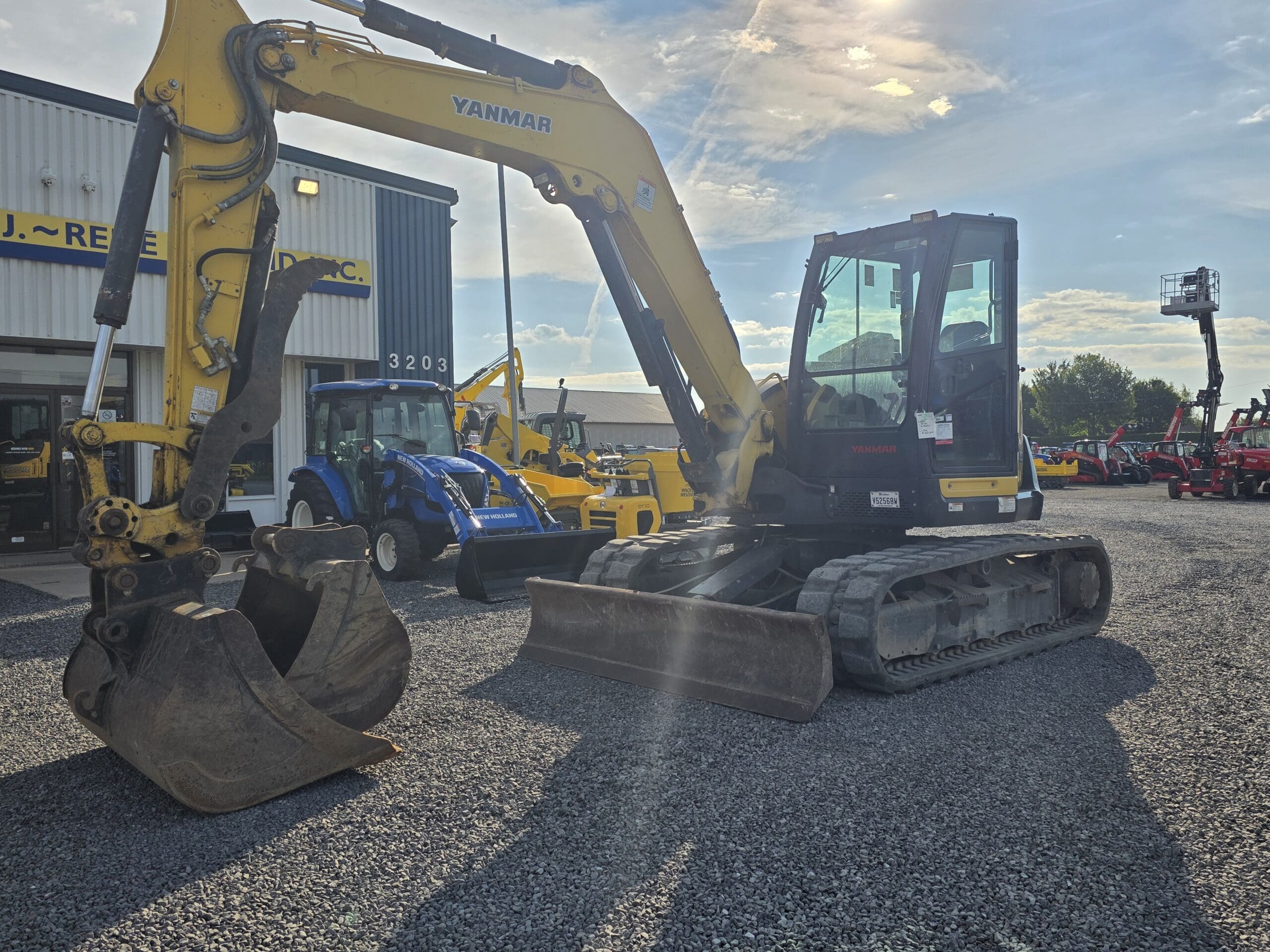 Une pelleteuse Yanmar jaune à chenilles en caoutchouc est garée sur un terrain en gravier à l'extérieur d'un concessionnaire. D'autres équipements de construction et agricoles sont visibles en arrière-plan sous un ciel partiellement nuageux.