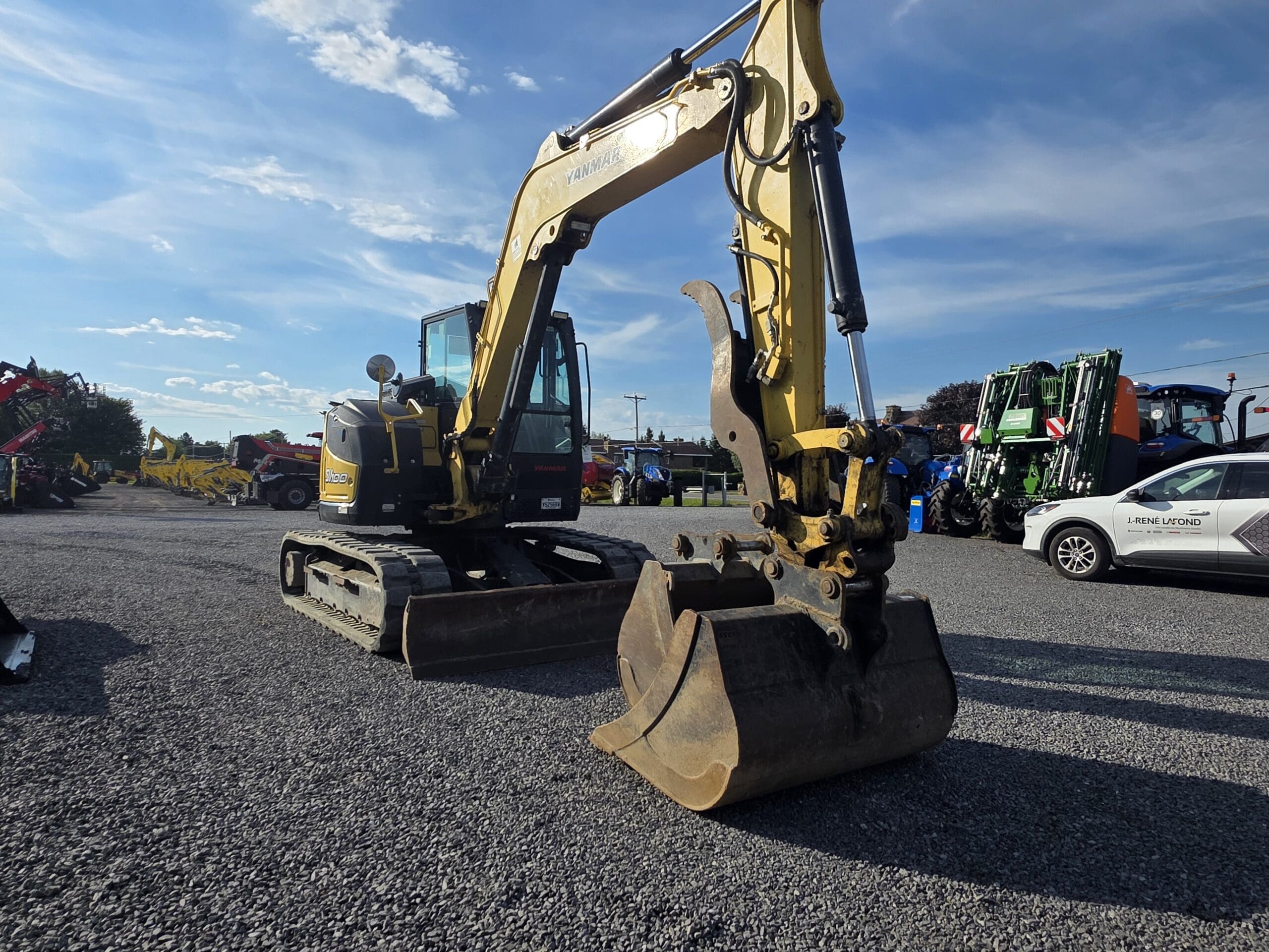Une pelleteuse jaune équipée d'un grand godet est garée sur du gravier dans un terrain avec d'autres machines et une voiture blanche. Le ciel est bleu avec des nuages épars.