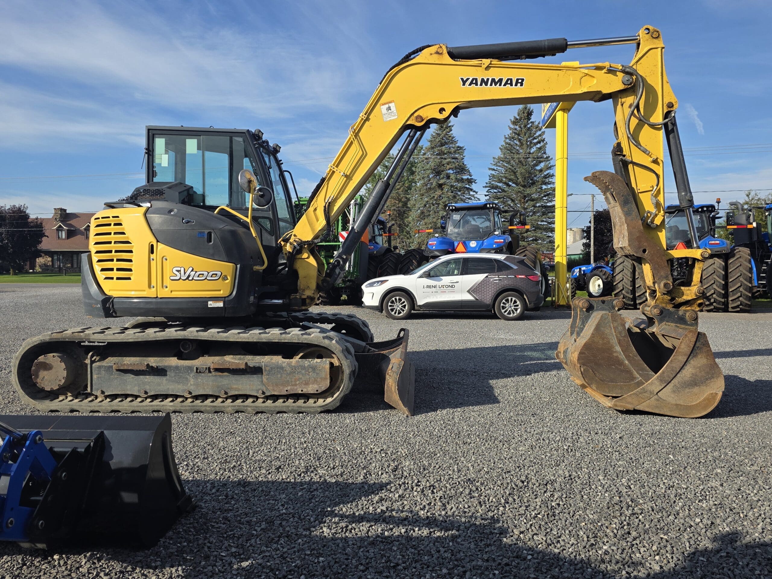 Une pelleteuse jaune Yanmar SV100 équipée d'une grande griffe est garée sur du gravier à côté d'un SUV blanc et de plusieurs tracteurs bleus, avec des arbres et un ciel bleu en arrière-plan.