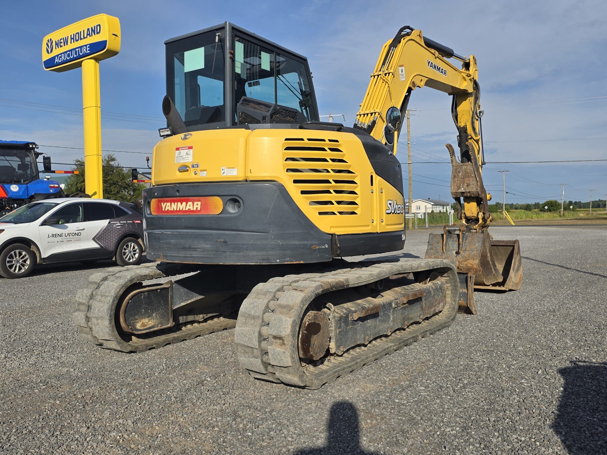 Une pelle compacte Yanmar jaune avec des chenilles en caoutchouc est garée sur du gravier près d'un panneau New Holland Agriculture et de véhicules, par une journée ensoleillée avec un ciel clair.