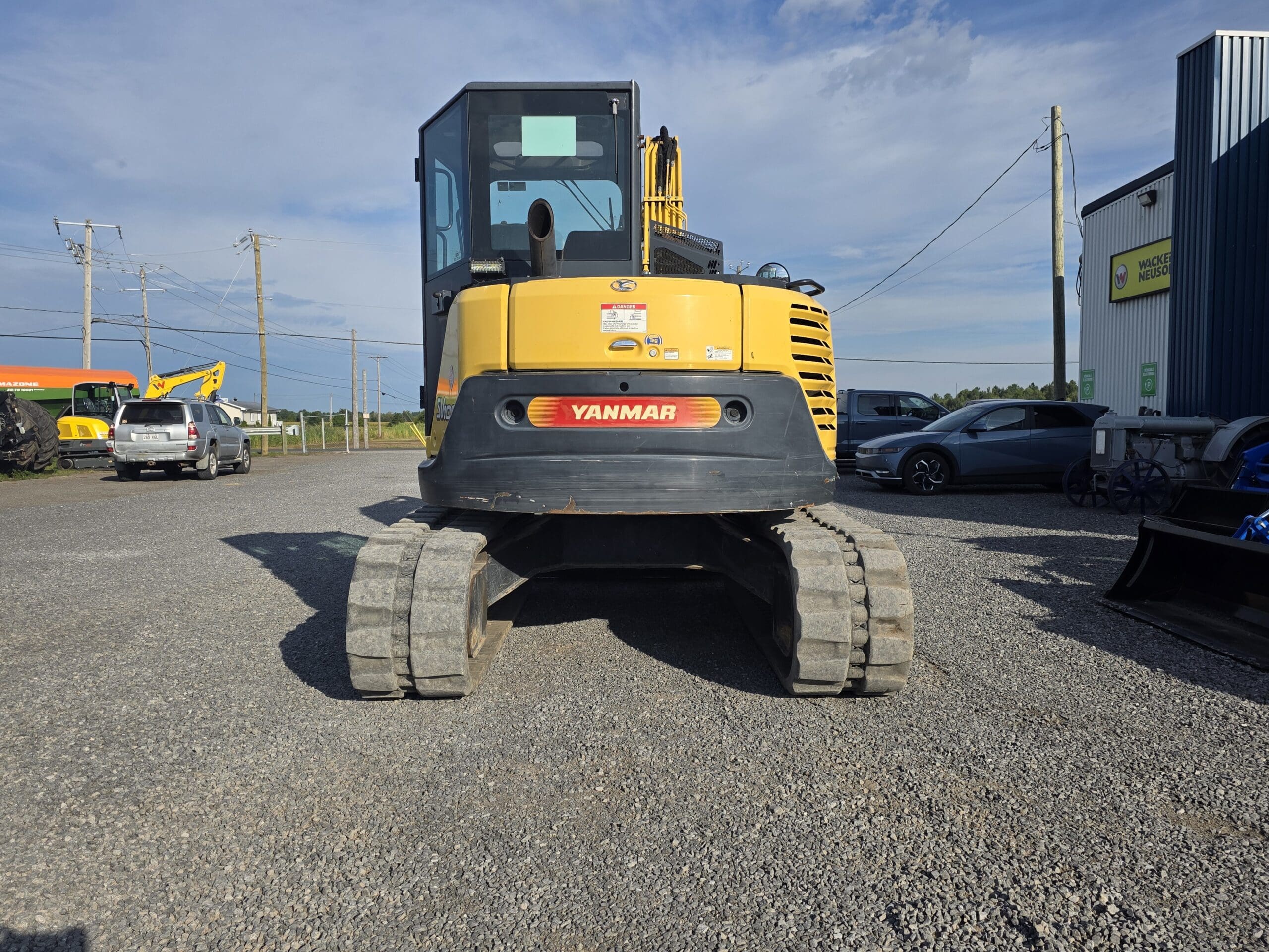 Une pelleteuse Yanmar jaune à chenilles en caoutchouc est garée sur un terrain en gravier sous un ciel bleu, avec des voitures et des bâtiments visibles à l'arrière-plan.