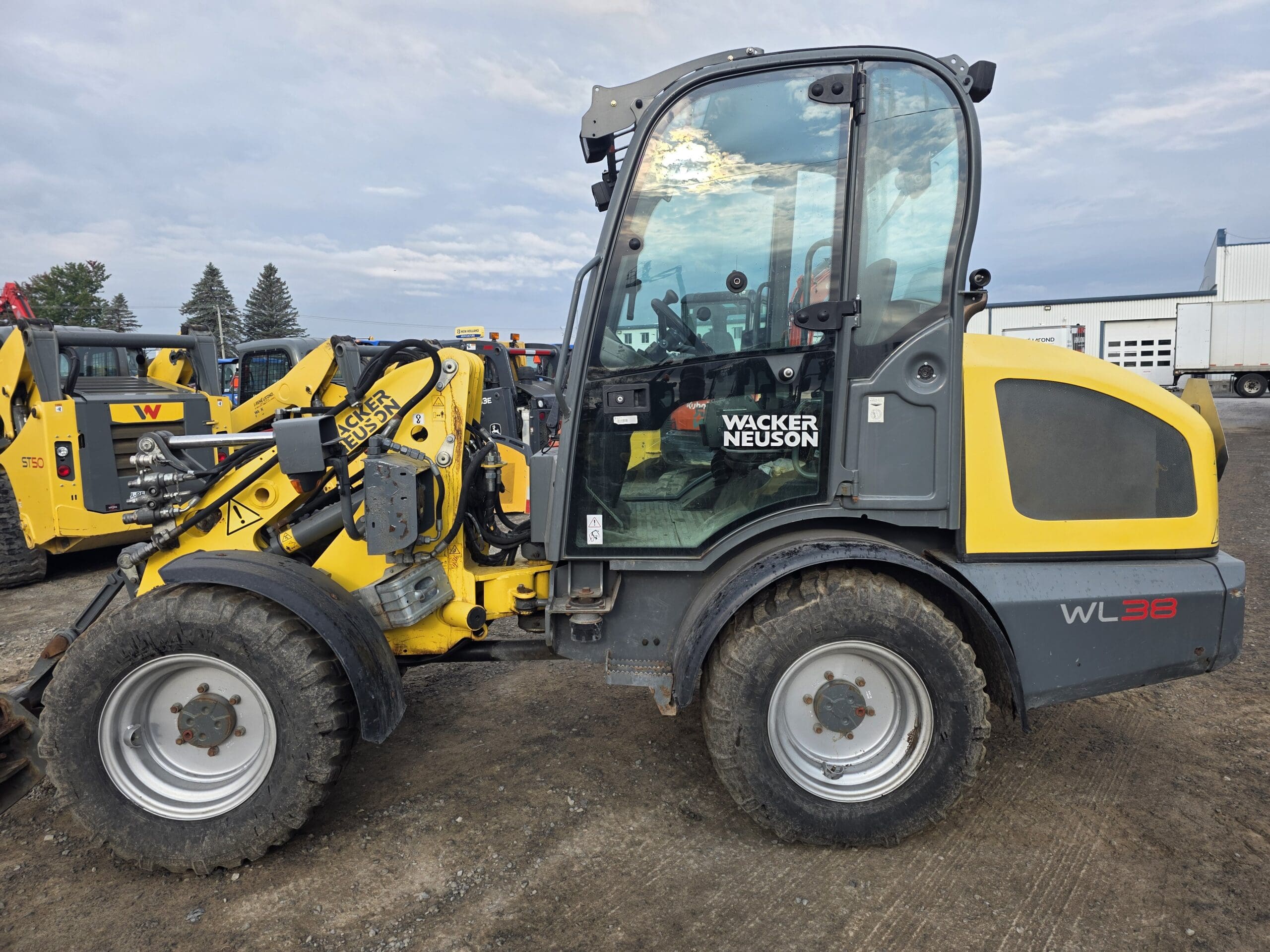 Une chargeuse sur pneus Wacker Neuson WL38 jaune et grise est garée sur un terrain en gravier. D'autres machines et bâtiments sont visibles en arrière-plan sous un ciel nuageux.