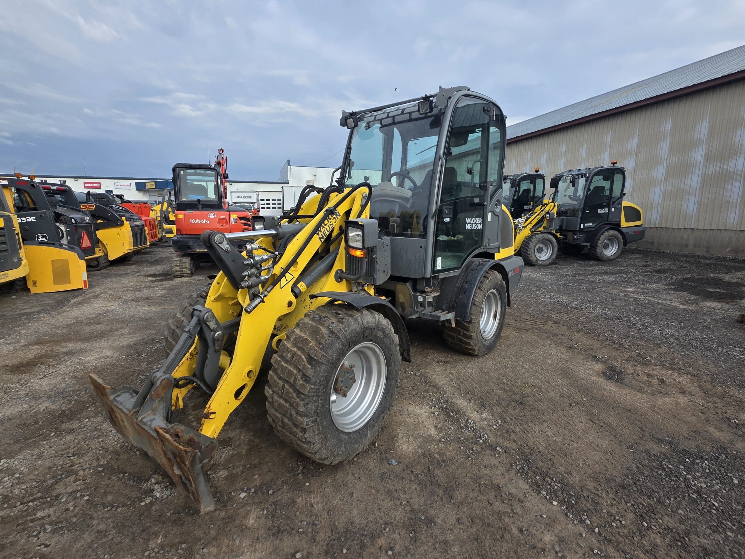 Un véhicule de construction à chargeur frontal jaune et noir est garé sur un terrain en gravier. D'autres machines et équipements similaires sont visibles à l'arrière-plan, près d'un grand bâtiment métallique.