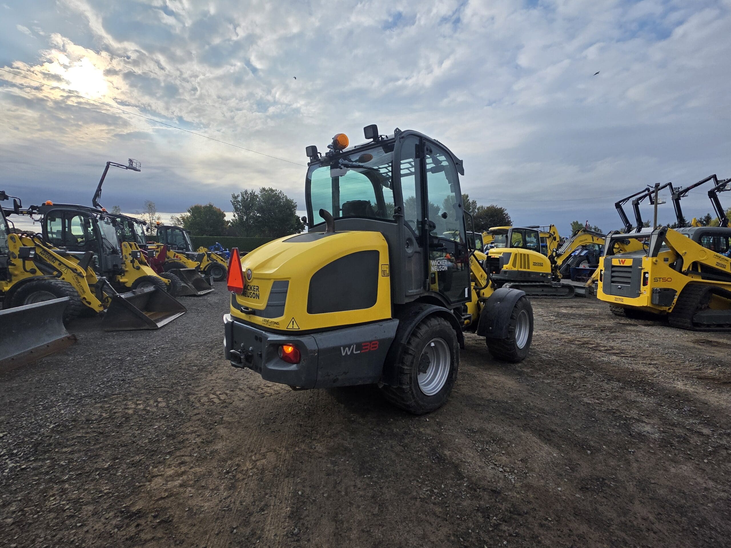 Une chargeuse sur pneus WL38 jaune est garée sur du gravier dans un parc de matériel de construction, entourée d'autres chargeuses et machines jaunes, sous un ciel nuageux où le soleil est partiellement visible.