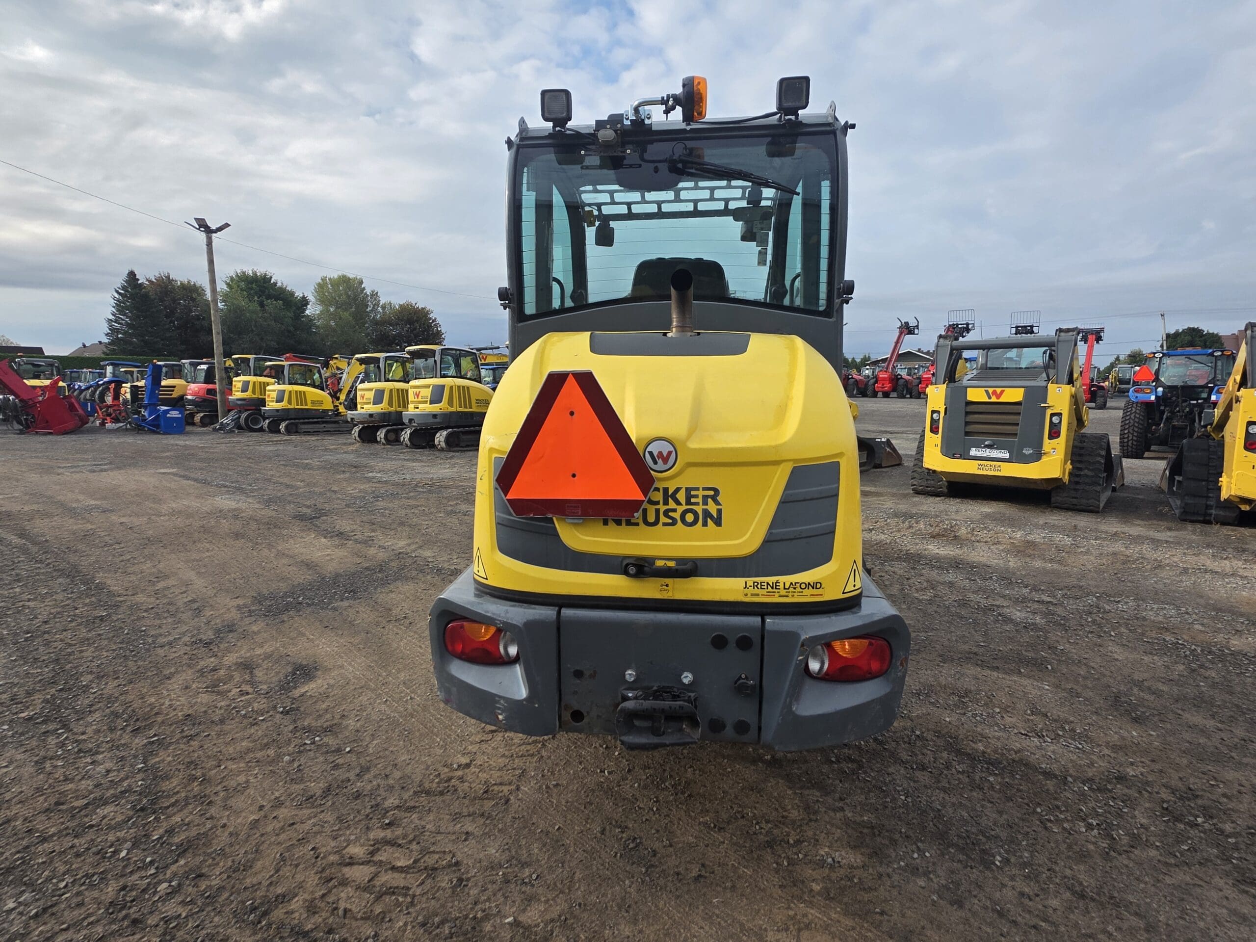 Un véhicule de chantier Wacker Neuson jaune avec un panneau triangulaire orange de signalisation de véhicule lent à l'arrière, garé sur un terrain en gravier avec d'autres machines à l'arrière-plan.