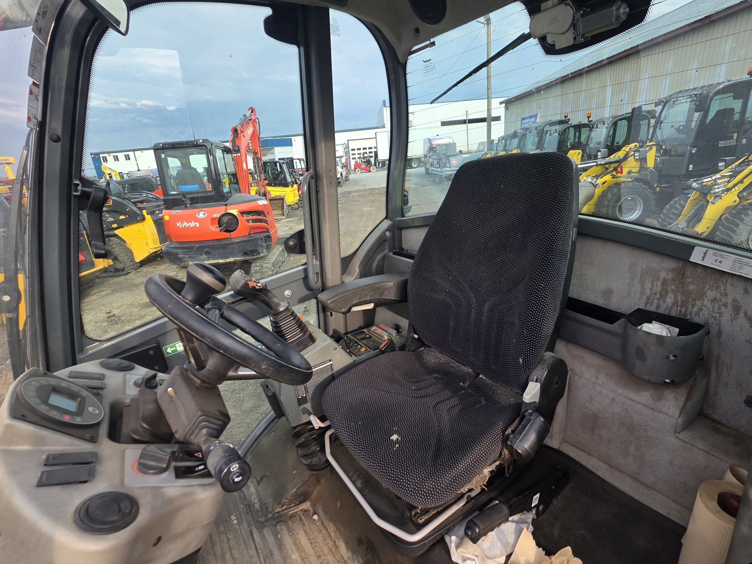 Vue de l'intérieur de la cabine d'un véhicule de construction montrant le siège du conducteur, le volant et les panneaux de commande. Diverses machines de construction sont visibles à l'extérieur par la fenêtre.