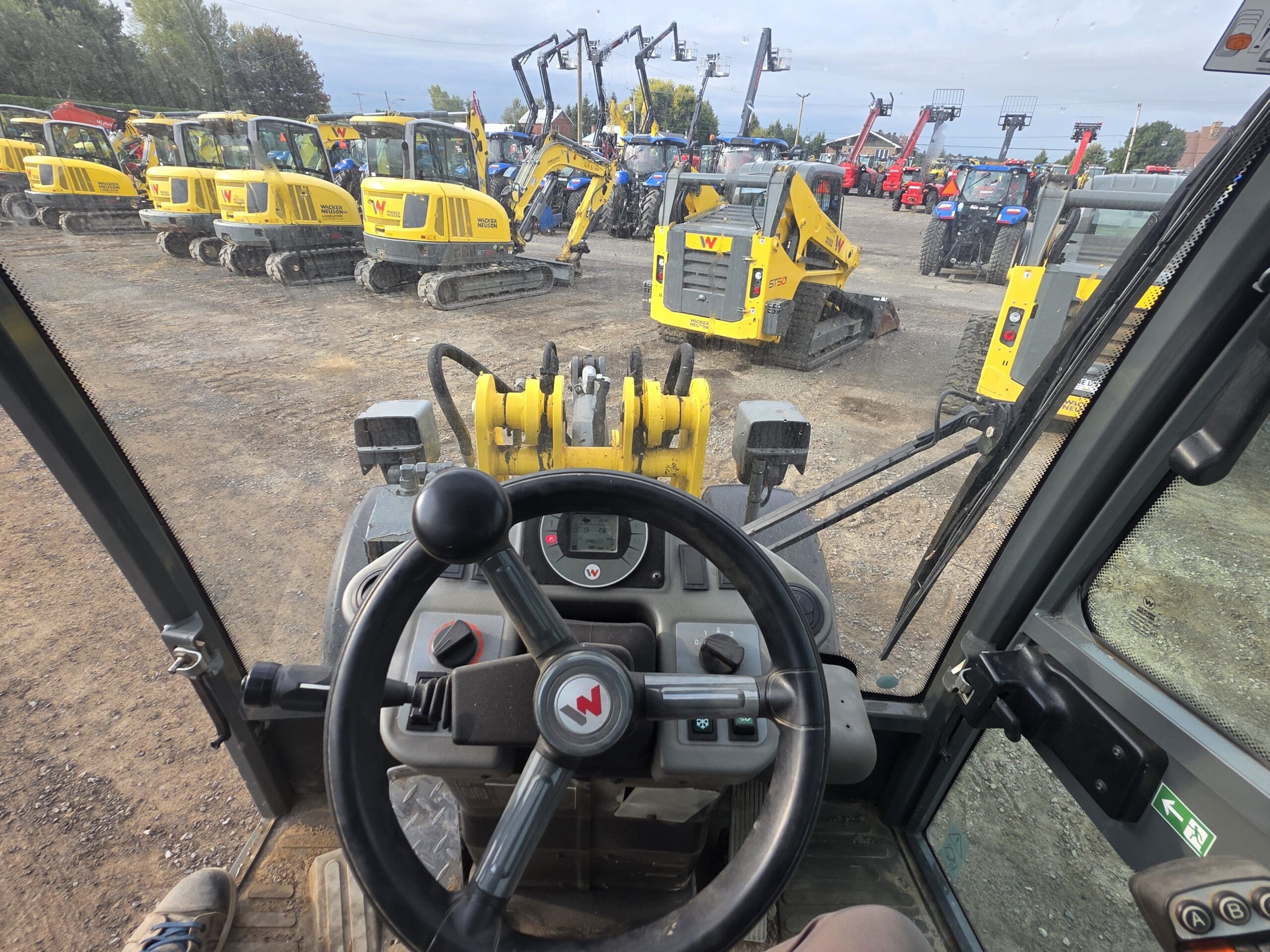 Vue de l'intérieur de la cabine d'un véhicule de construction, montrant le volant, les commandes et le tableau de bord, avec plusieurs excavatrices jaunes et des équipements de construction garés à l'extérieur sur un terrain en gravier.