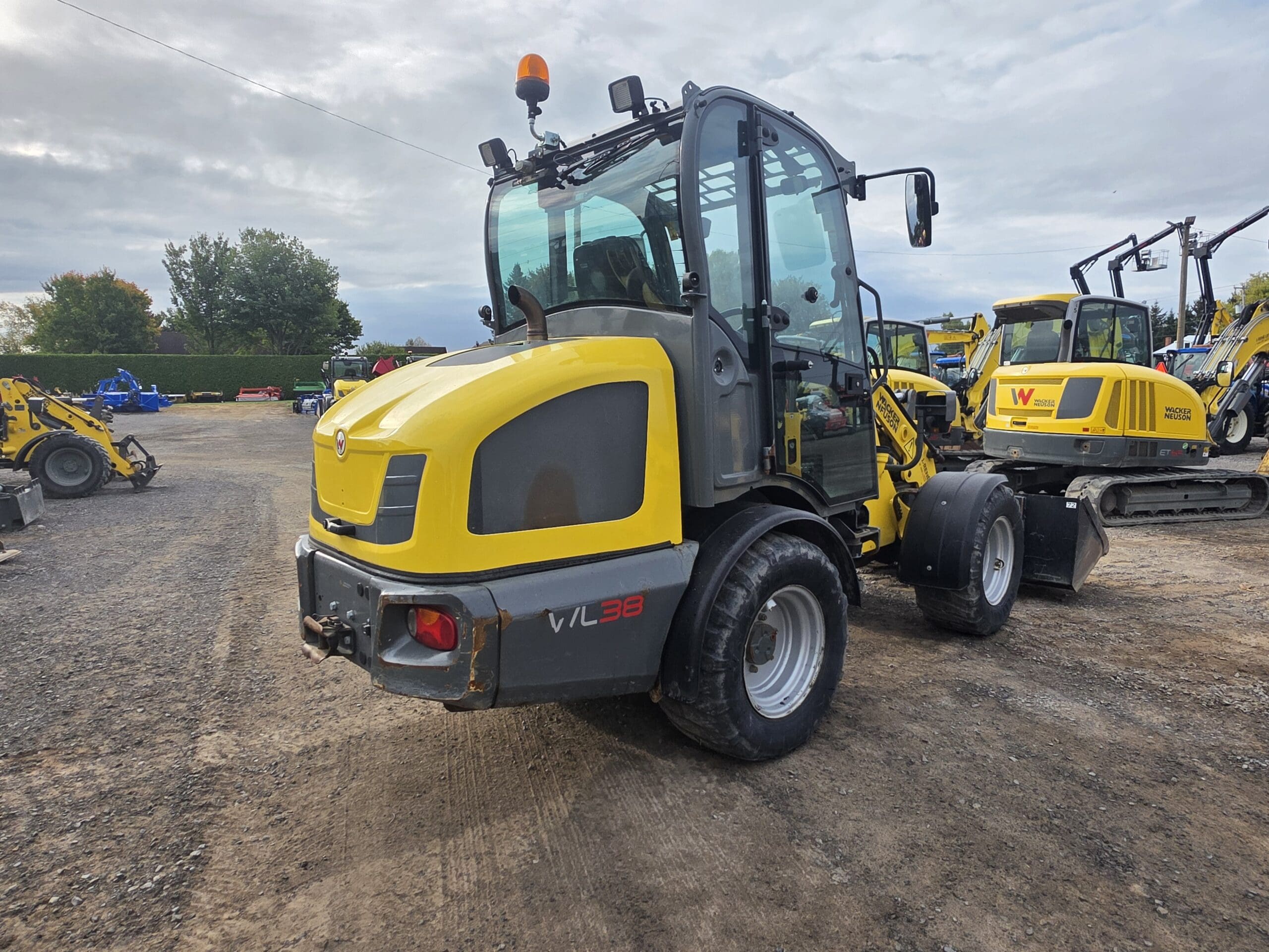 Un véhicule de construction jaune et gris, probablement une chargeuse compacte sur roues, est garé sur un terrain en gravier avec d'autres machines similaires en arrière-plan sous un ciel nuageux.