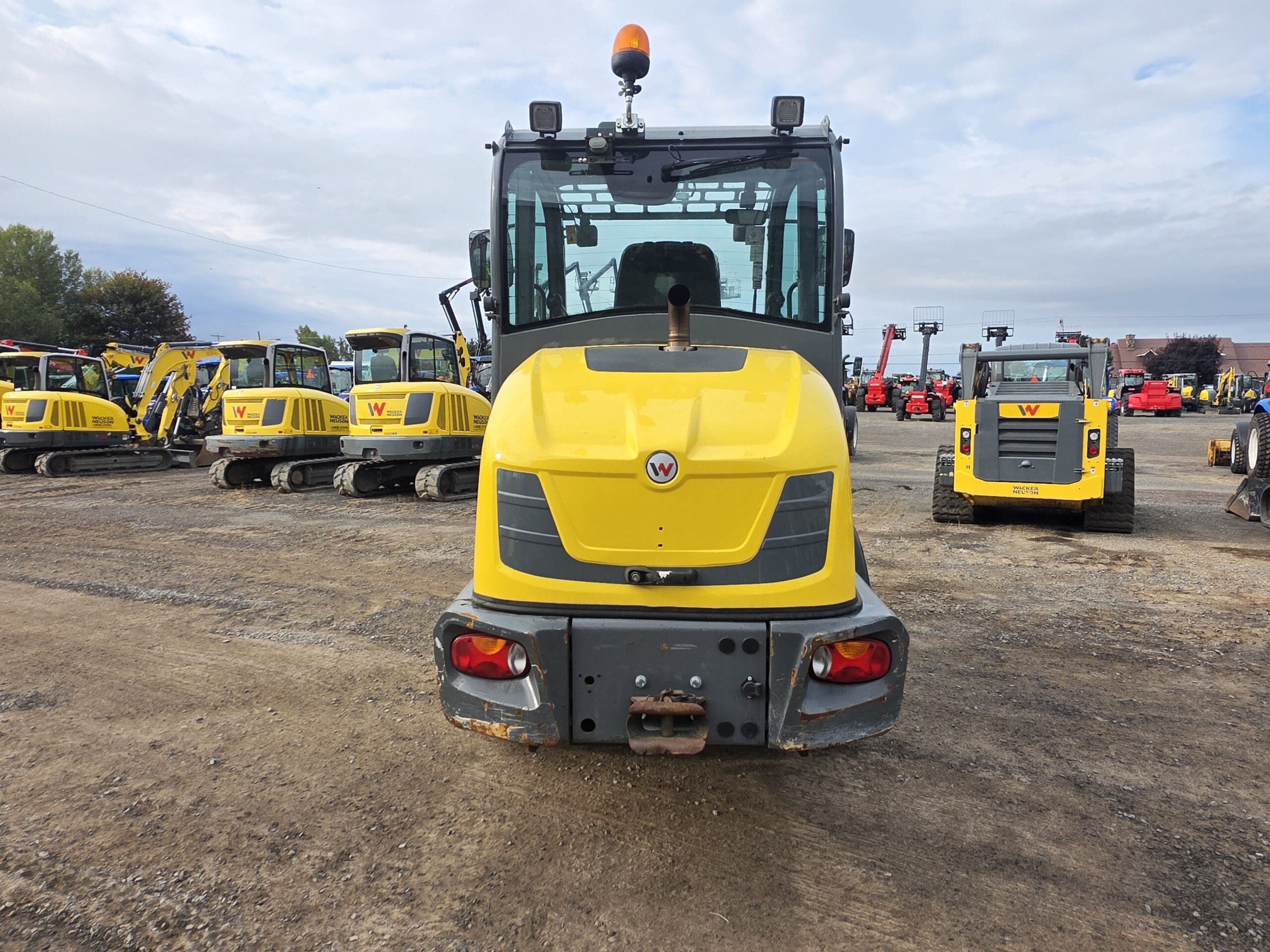Un véhicule de chantier jaune vu de l'arrière, garé sur un terrain avec d'autres machines similaires sous un ciel nuageux.