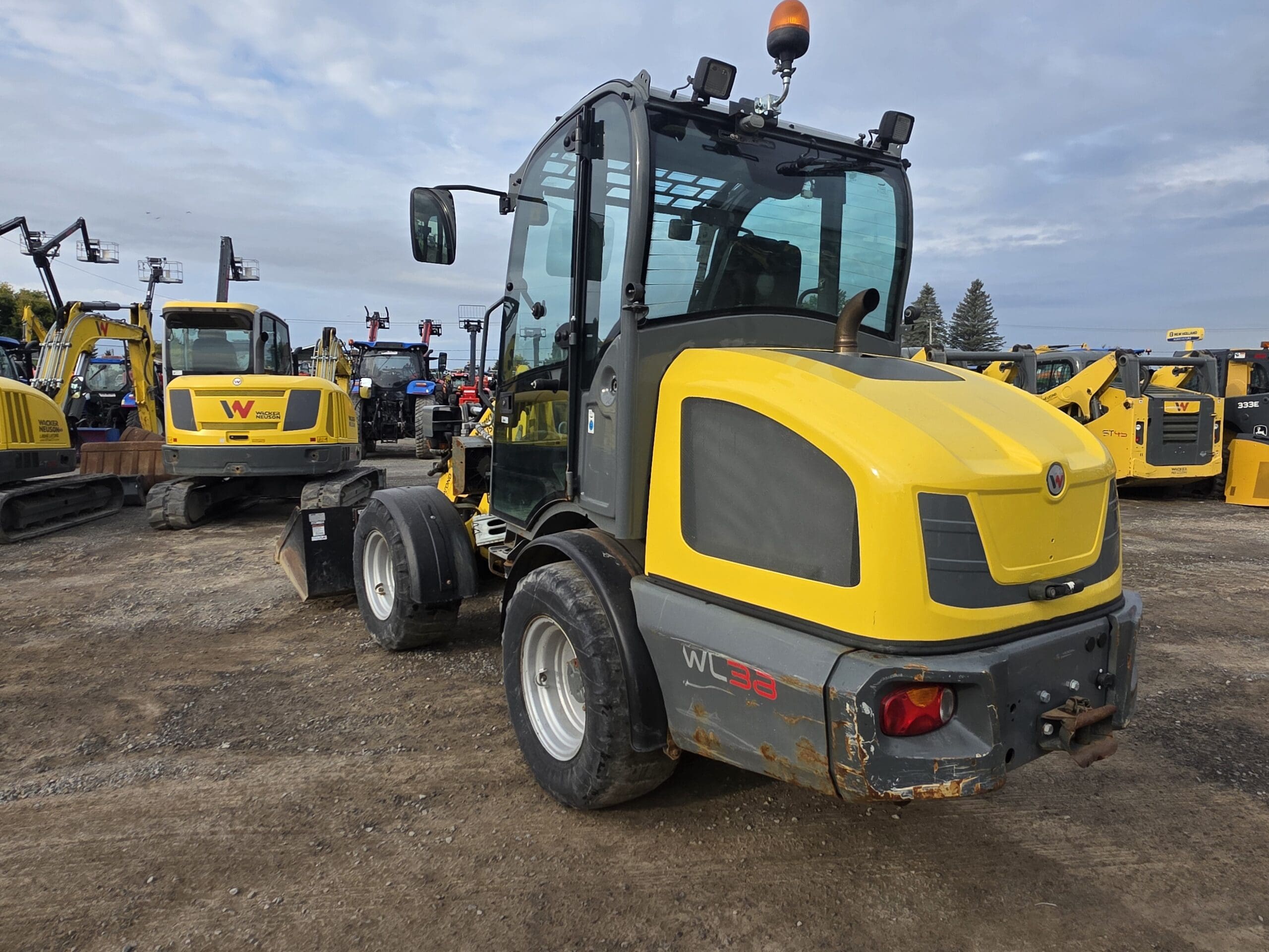 Une chargeuse compacte sur pneus jaune et grise est garée sur un terrain en terre battue parmi d'autres véhicules et équipements de construction, sous un ciel nuageux.