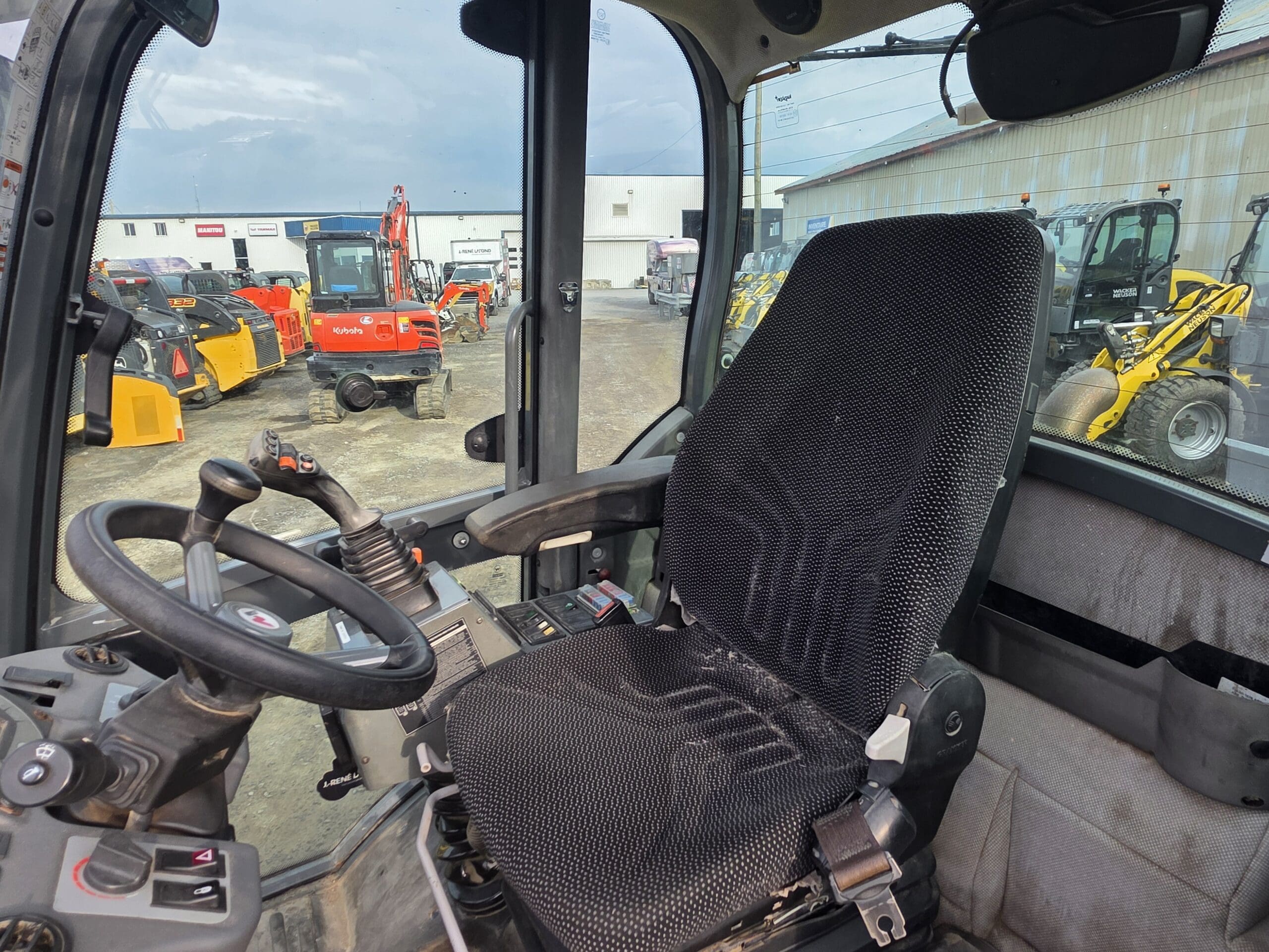 Vue de l'intérieur de la cabine d'un véhicule de construction, montrant le siège du conducteur, le volant et le panneau de commande. À l'extérieur, divers engins et équipements de construction sont visibles dans une cour.