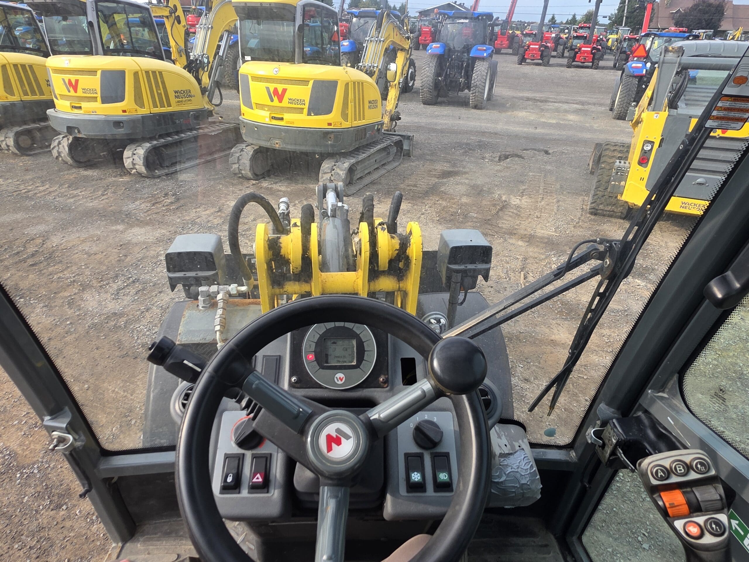 Vue de l'intérieur de la cabine d'un véhicule de construction, montrant le volant, le tableau de bord et les commandes, avec plusieurs excavatrices jaunes et tracteurs rouges garés à l'extérieur sur un terrain en gravier.