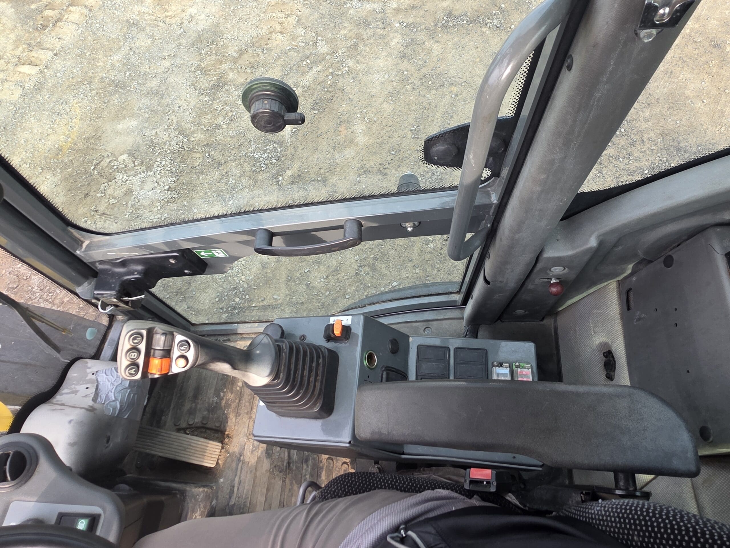 Vue de l'intérieur d'une cabine de véhicule de chantier, montrant le levier de commande, les interrupteurs, les pédales, l'accoudoir et la porte, avec une surface de gravier visible à l'extérieur par la fenêtre.