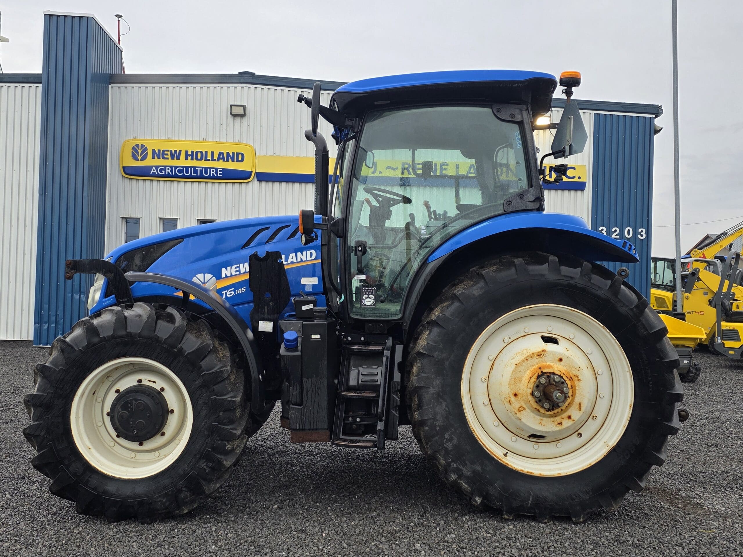 Un tracteur bleu New Holland T6.160 est garé sur du gravier devant le bâtiment d'un concessionnaire New Holland Agriculture.