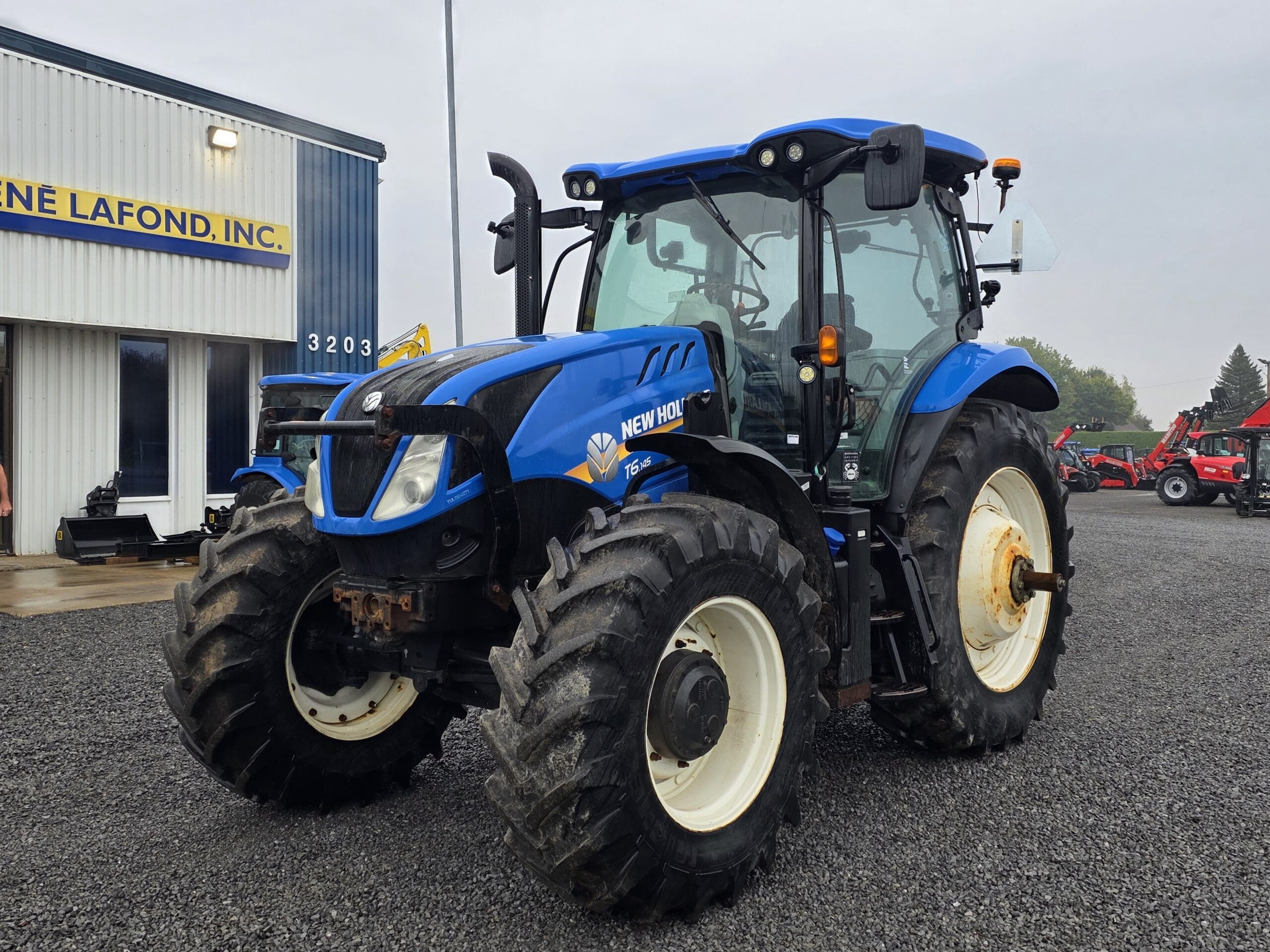 Un tracteur New Holland bleu garé sur du gravier devant un bâtiment sur lequel on peut lire 