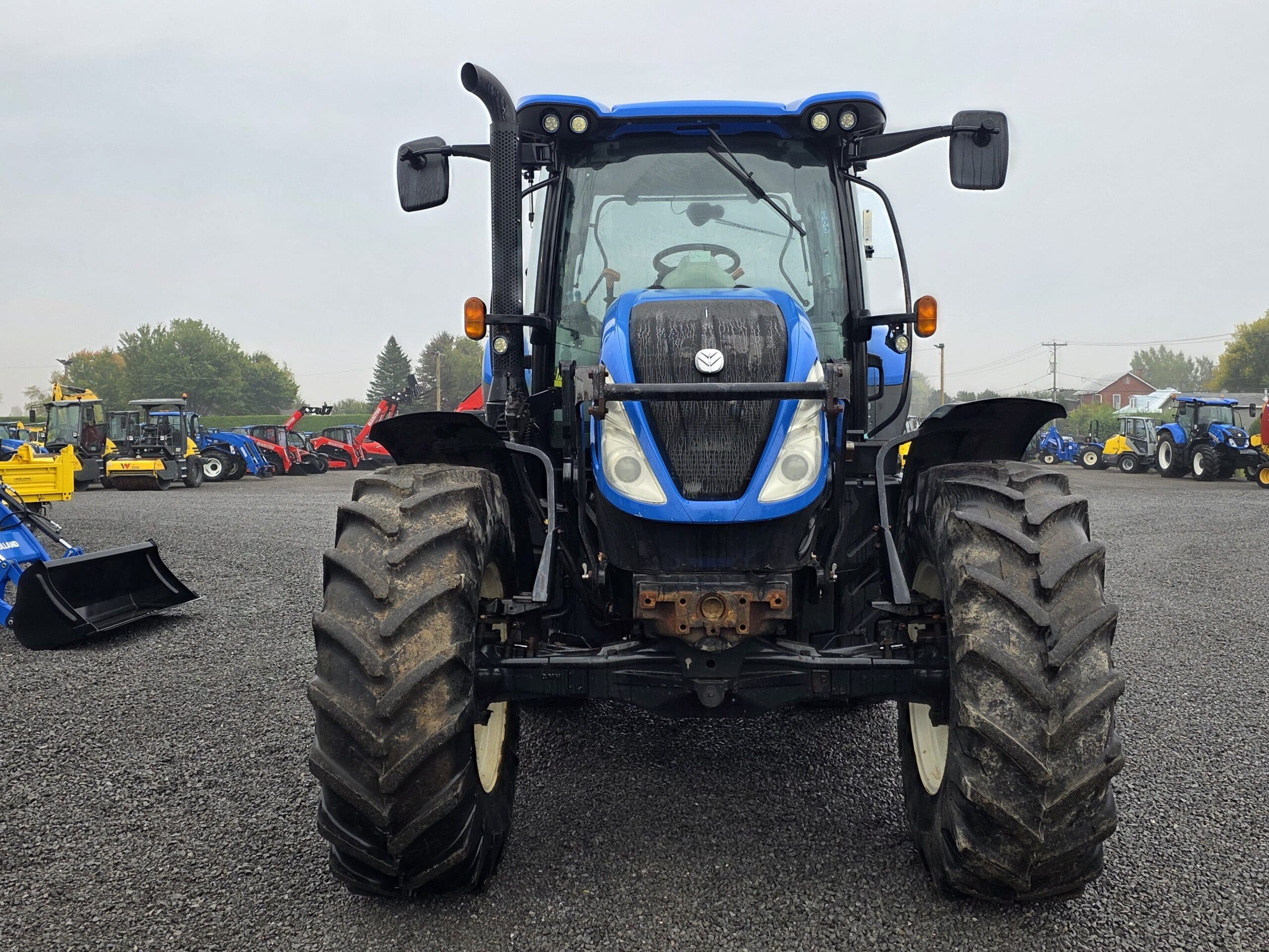 Un tracteur bleu vu de l'avant, garé sur un terrain en gravier avec d'autres tracteurs et équipements en arrière-plan sous un ciel nuageux.