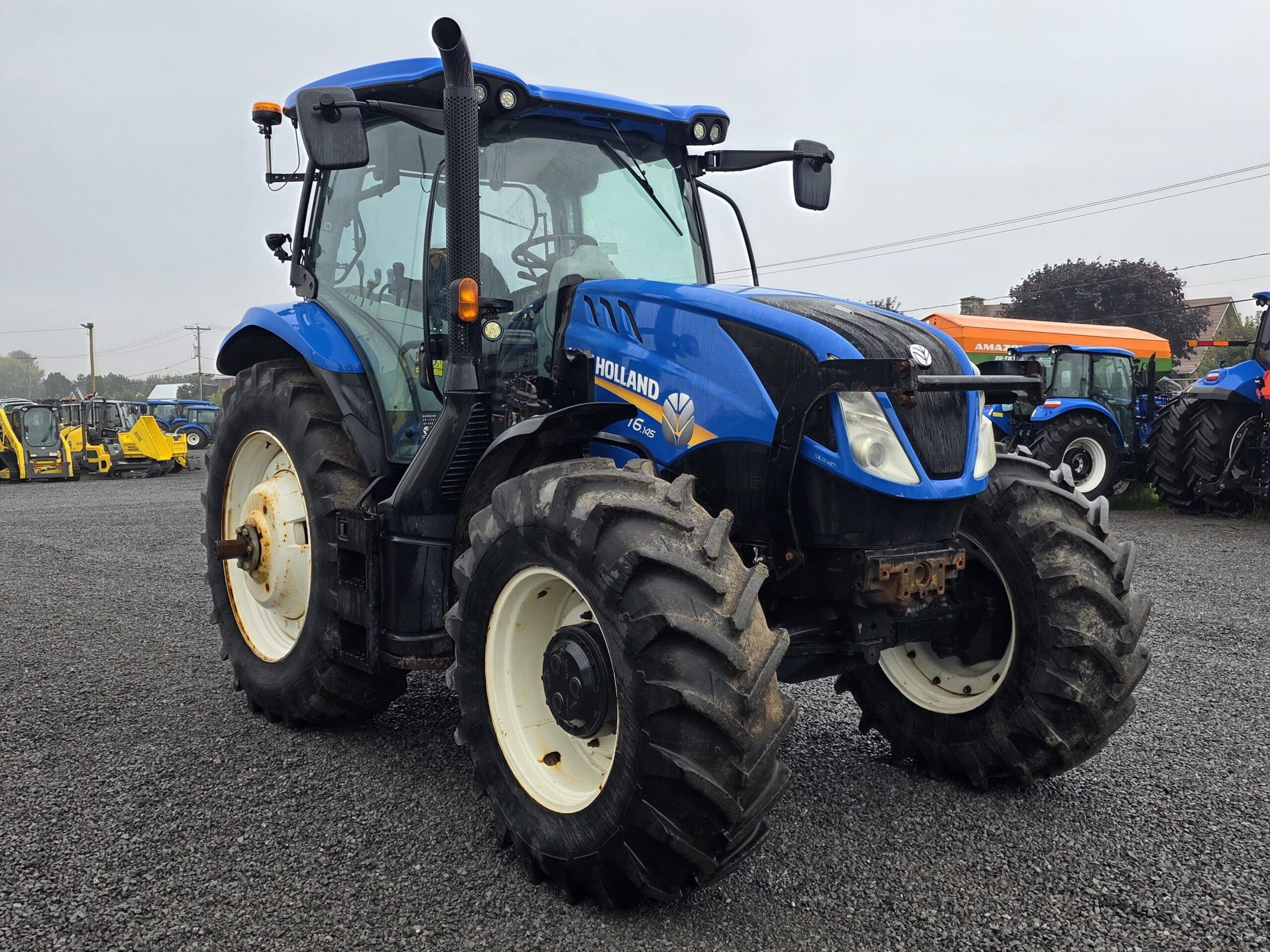 Par une journée nuageuse, un tracteur agricole New Holland bleu aux pneus larges et robustes est garé sur un terrain en gravier parmi d'autres machines.